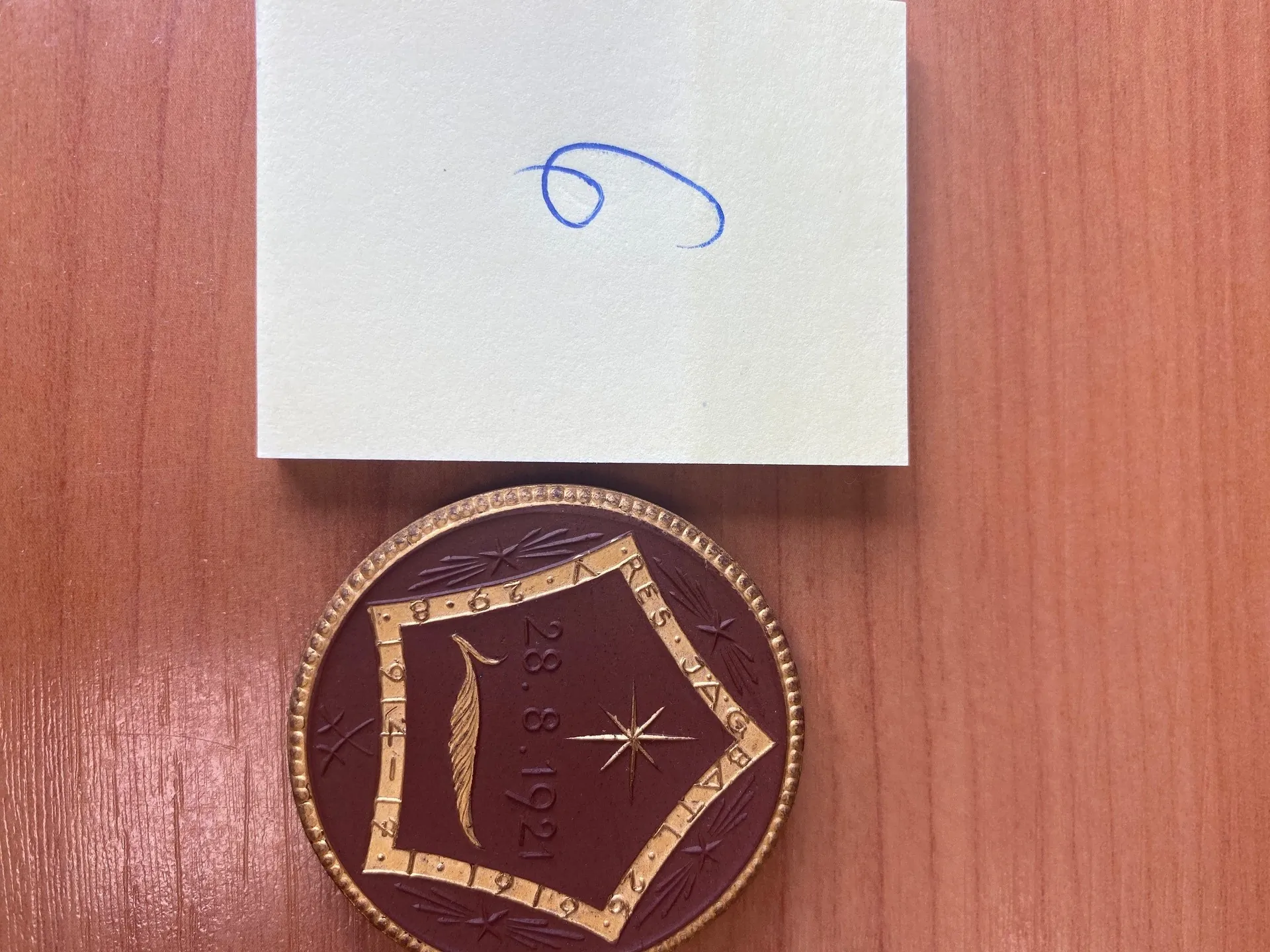Na drewnianym tle o poziomych liniiach znajdują się biały kartonik z niebieskim, ręcznym znakiem oraz okrągły emblemat. Emblemat ma brązowe tło z złotymi detalami: w centralnej części romb z napisem "28.8.1921", piórem, gwiazdą i napisem "RES JAG" na obramowaniu. Wokół rombu widoczne są ozdobne elementy i cyfry "1921".