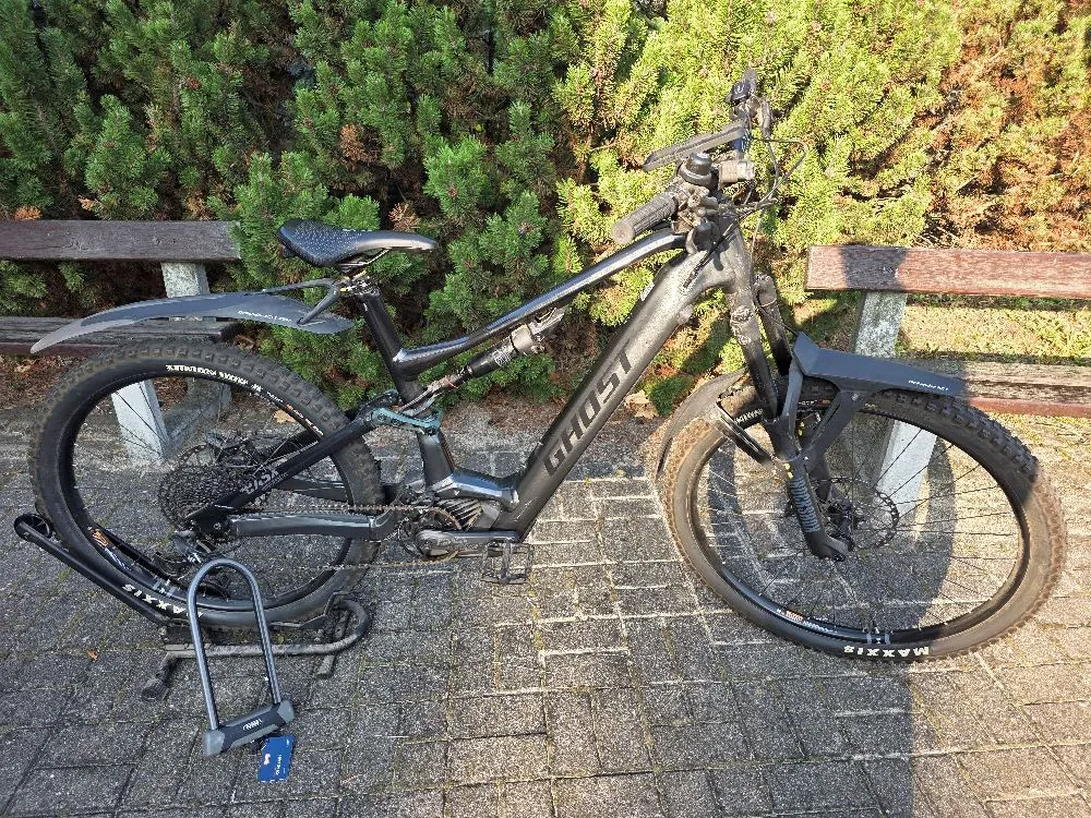 Czarny rower elektryczny marki Ghost z napisem GHOST na ramie, zaparkowany na kostce brukowej przed drewnianą ławką i zielonymi krzewami. Rower posiada amortyzator przedni i tylny, szerokie opony Maxxis oraz błotniki.