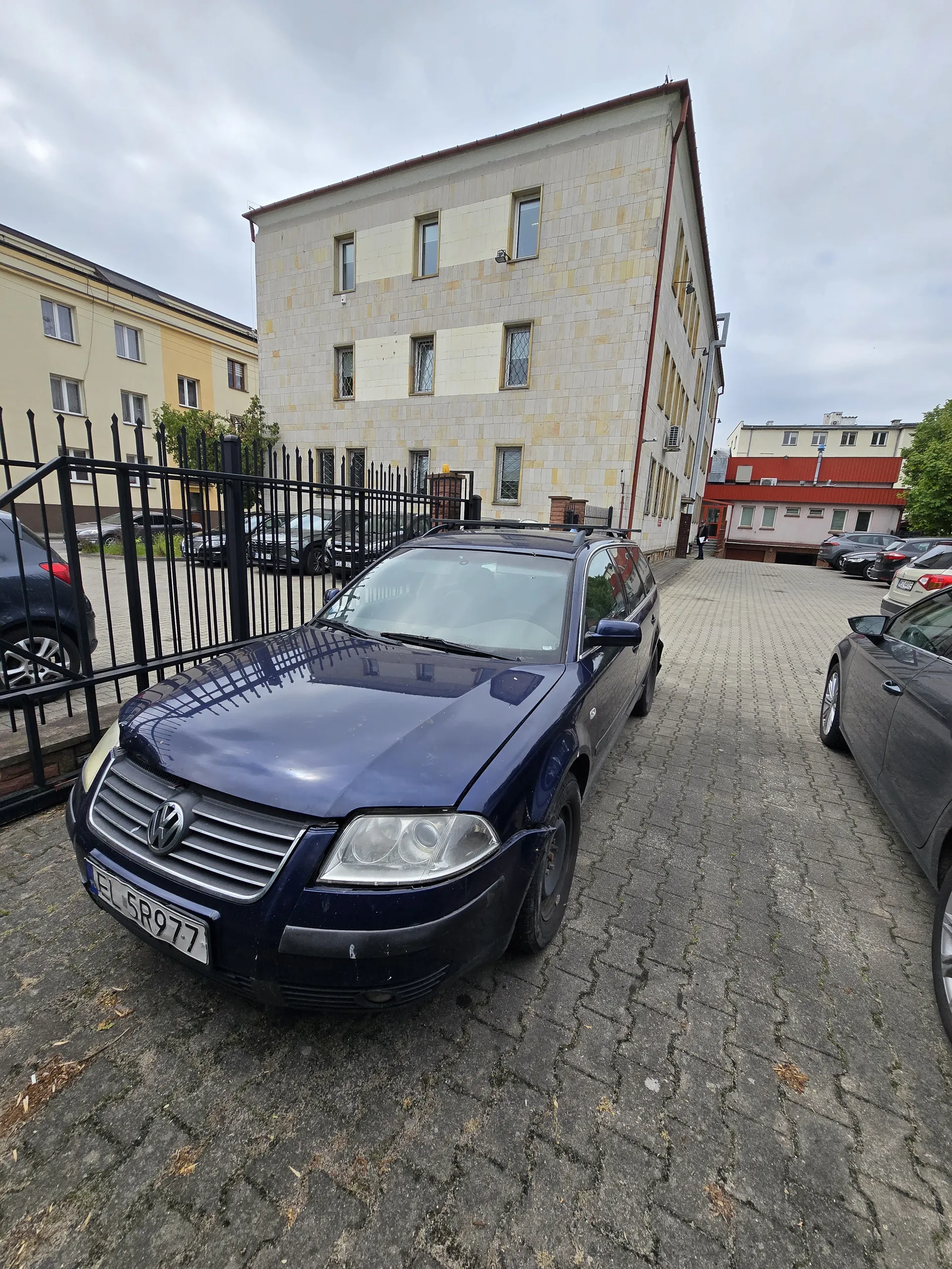 Ciemnoniebieski samochód marki Volkswagen zaparkowany na brukowanej powierzchni. Tablica rejestracyjna EL 5R977. Za samochodem czarne metalowe ogrodzenie. W tle widoczne są wielopiętrowe budynki o jasnych elewacjach z oknami. Niebo jest zachmurzone. W oddali zaparkowane są inne samochody. Na pierwszym planie fragment brukowanej nawierzchni i drzewo.