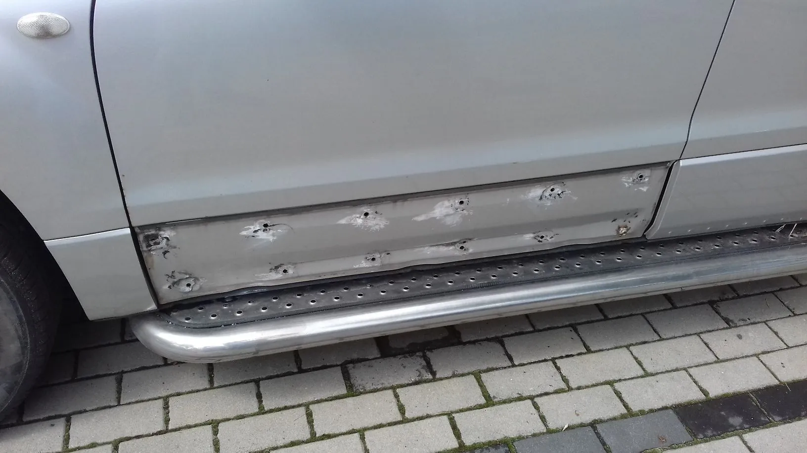 Zbliżenie na dolną część srebrnego samochodu. Widoczny jest próg z metalowym stopniem, nad którym znajduje się uszkodzony element karoserii z licznymi wgnieceniami i śladami po naprawach szpachlowych. Samochód stoi na kostce brukowej.