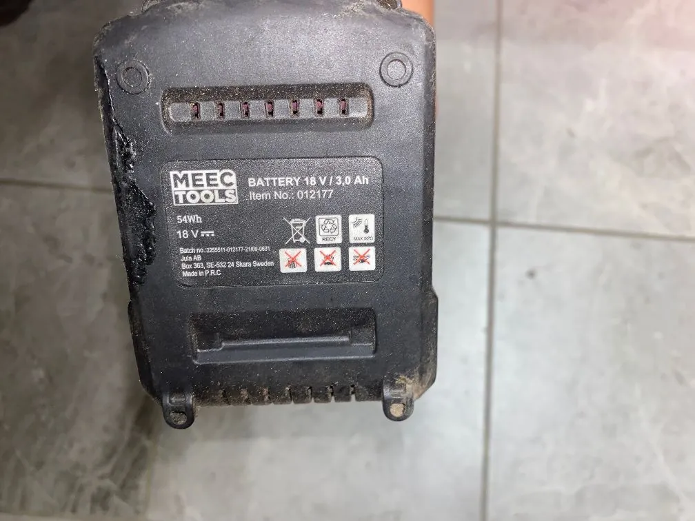 Zbliżenie na czarną, plastikową obudowę akumulatora marki MEEC TOOLS. Na obudowie znajduje się srebrna etykieta z tekstem: 'MEEC TOOLS BATTERY 18 V / 3,0 Ah Item No.: 012177'. Poniżej widnieją oznaczenia '54Wh' oraz '18 V', a także symbole ostrzegawcze dotyczące utylizacji i bezpieczeństwa. Na dole etykiety znajduje się tekst: 'Batch no.: 2205511-01-012177-21/08-0631', 'AB MEEC Box 363, SE-532 24 Skara Sweden Made in P.R.C'. Obudowa jest zakurzona i posiada widoczne ślady użytkowania.