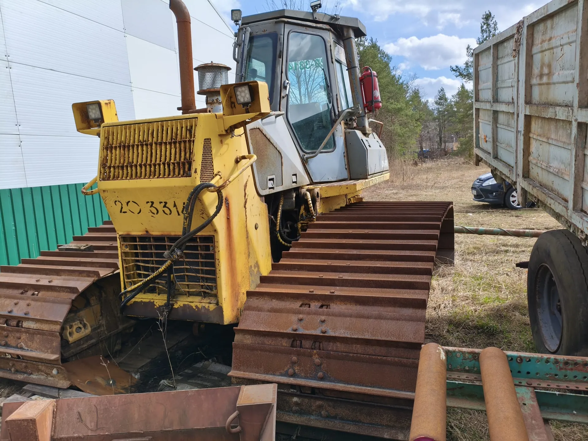 Żółty pojazd budowlany z gąsienicami, oznaczony numerem 20331, z widocznym tekstem A188-IX na szybie kabiny. Pojazd ma rdzewne gąsienice i elementy metalowe. W tle biały budynek z zielonym pasem, rdzawa przyczepa oraz częściowo widoczny czarny samochód. Niebo z chmurami.