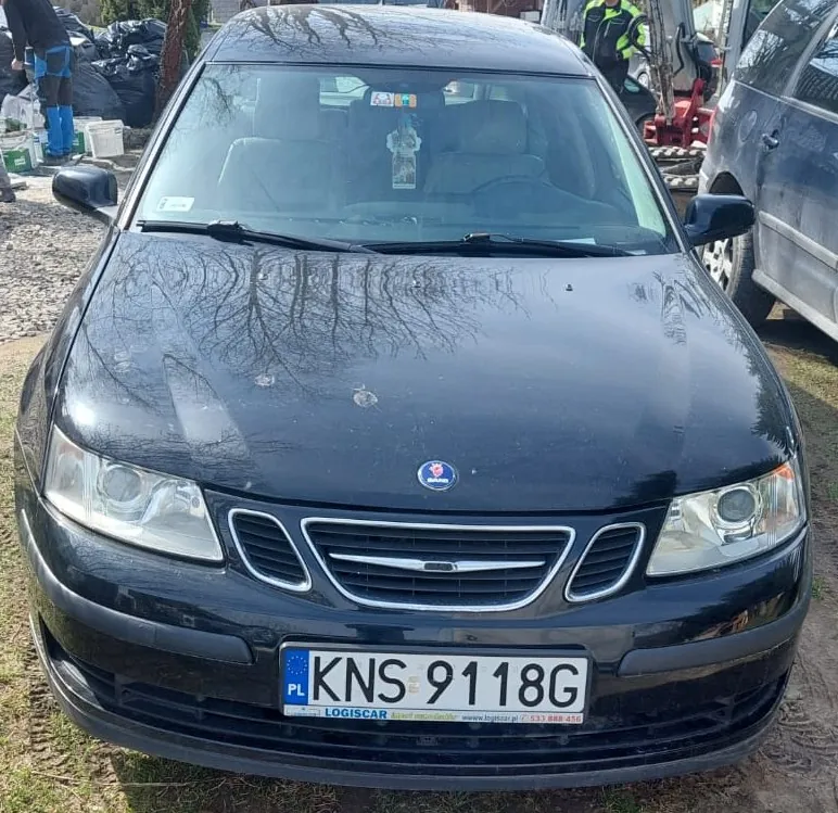 Czarny samochód osobowy marki Saab widziany od przodu. Pojazd ma tablicę rejestracyjną o numerze KNS 9118G z polskim oznaczeniem PL. Poniżej numeru rejestracyjnego znajduje się biała ramka z napisem LOGISCAR oraz adresem strony internetowej i numerem telefonu. Na masce samochodu widoczne są drobne uszkodzenia lakieru.