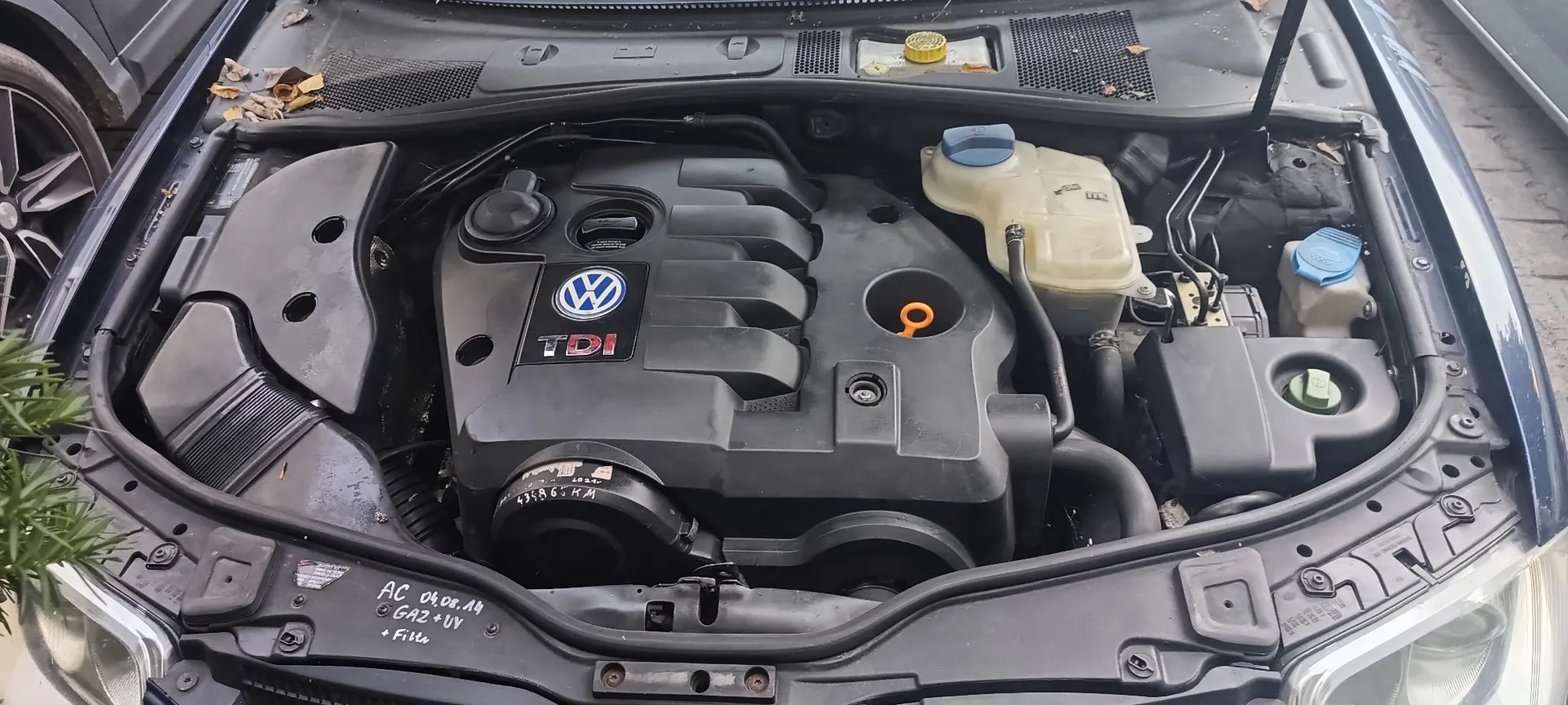 Widok z góry na komorę silnika samochodu marki Volkswagen. W centrum znajduje się czarna plastikowa osłona silnika z logo VW oraz napisem TDI. Po prawej stronie widoczne są zbiorniki z płynami eksploatacyjnymi z niebieskimi zakrętkami. W dolnej części zdjęcia, na ramie nadwozia, znajduje się odręczna notatka: 'AC 06.08.14 GAZ + UV + Filtry'.