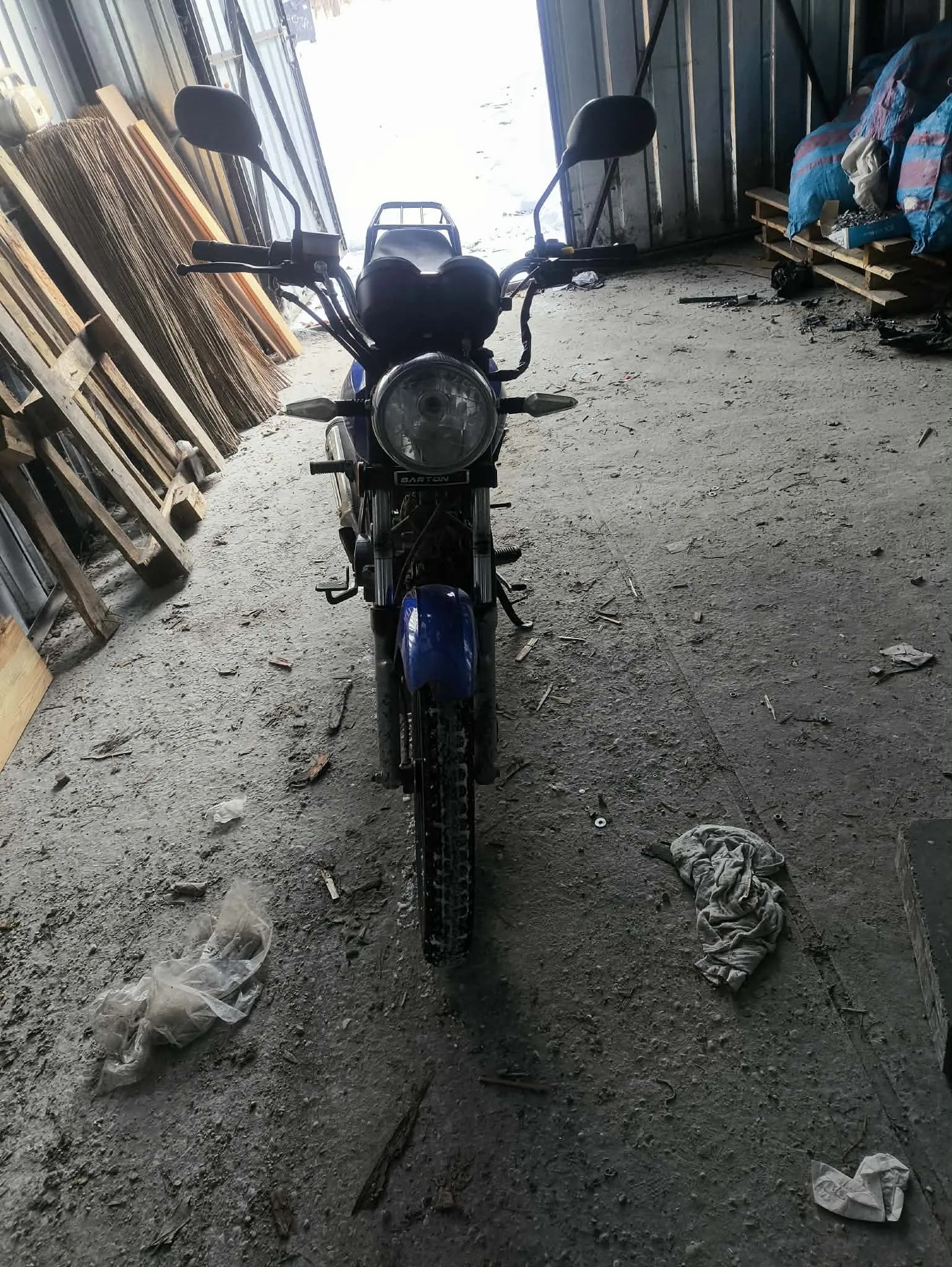Niebieski motocykl marki BARTON z przodem skierowanym do przodu, stojący w warsztacie. W tle widoczne są stosy desek po lewej stronie, palety z niebiesko-różowymi workami po prawej oraz rozrzucone na betonowej podłodze przedmioty, w tym plastikowa torba i szmatka. Na motocyklu widoczne lusterka, reflektor i czarna siedzisko.