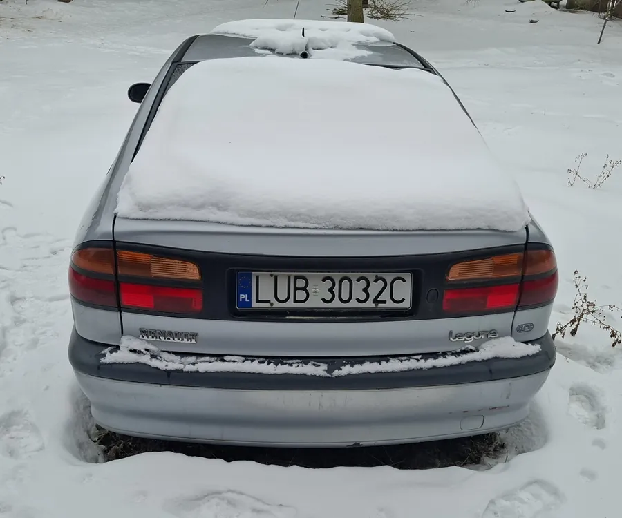 Tył srebrnego samochodu marki Renault Laguna pokrytego śniegiem. Na tablicy rejestracyjnej widoczny napis "LUB 3032C" oraz oznaczenie "PL". Samochód stoi na śnieżnej powierzchni z widocznymi śladami stóp. W tle białe tło z niewielkimi krzewami i drzewem.