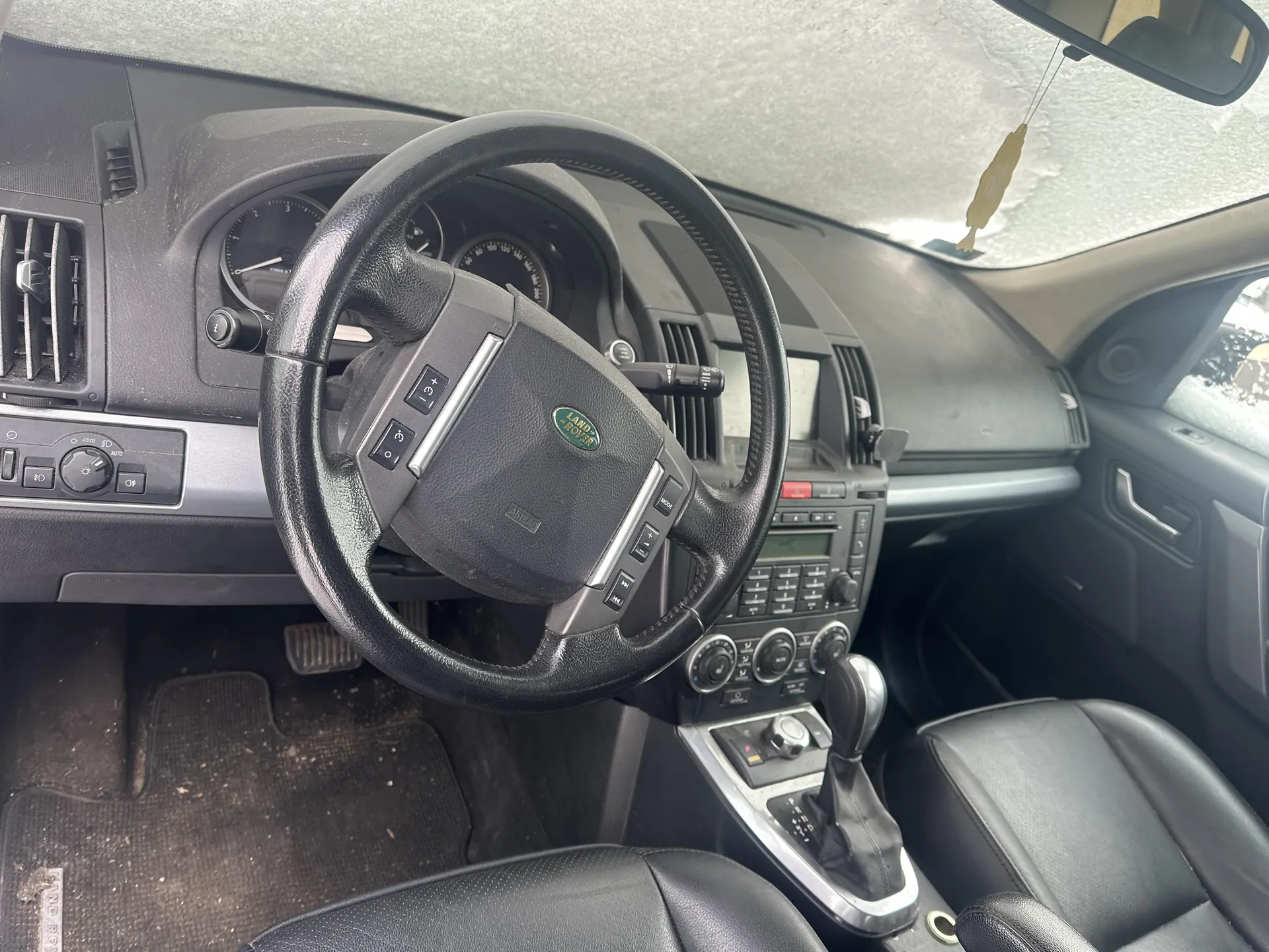 Wnętrze samochodu Land Rover z widocznym kierownicą z logo marki, czarnymi skórzonymi siedzeniami, deską rozdzielczą z licznikami, klimatyzacją i centralnym panelem sterowania. Na desce rozdzielczej znajdują się przyciski, pokrętła oraz wyświetlacz. W tle widać lusterko wsteczne z wiszącym żółtym aromatyzatorem. Podłoga pokryta jest brudnymi dywanikami. Kierownica posiada przyciski sterujące, a obok niej widoczny jest automatyczny selector biegów.