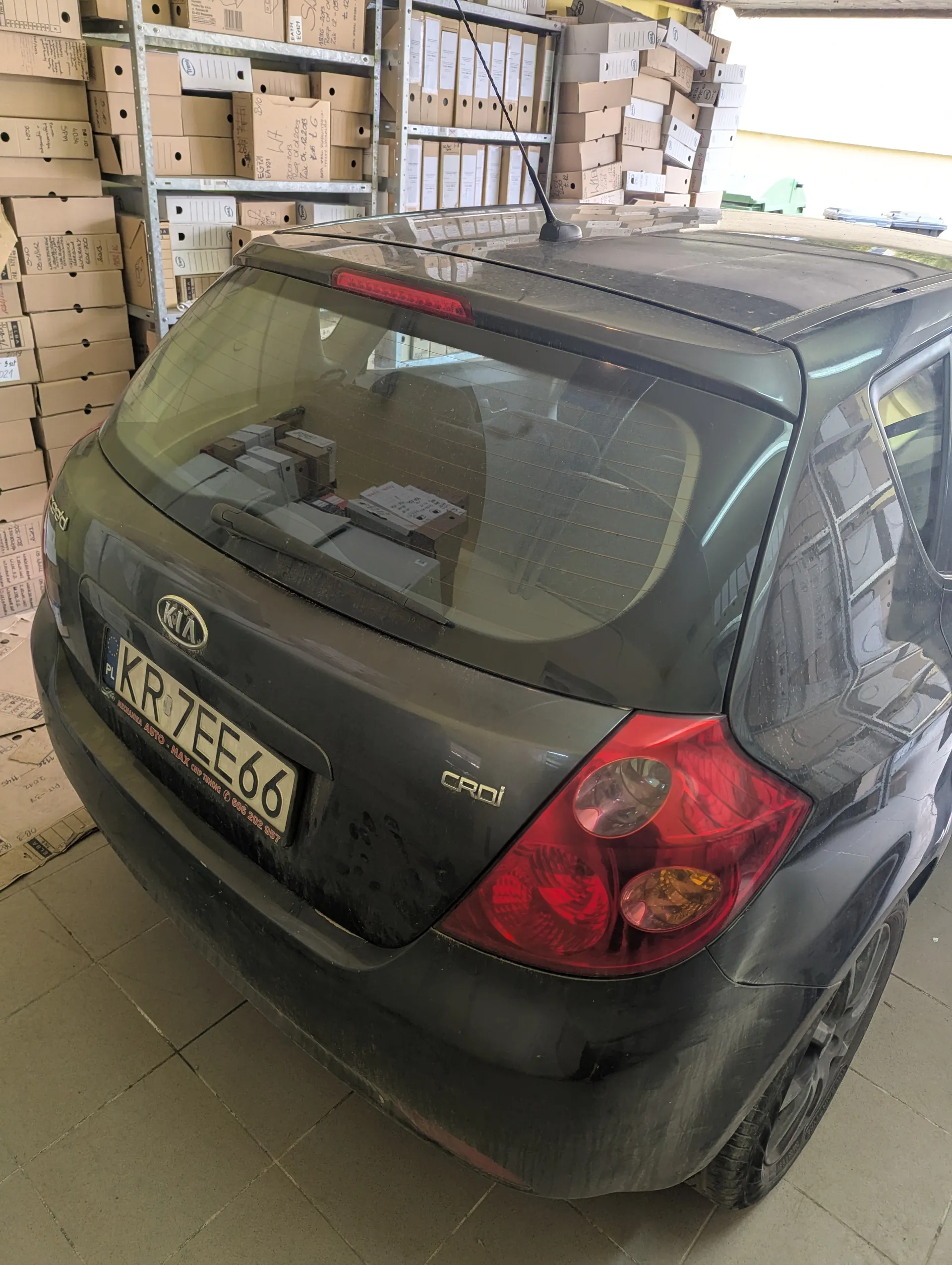 Z tyłu widoczny ciemnoszary samochód marki Kia model Ceed z tablicą rejestracyjną KR7EE66 (PL). Samochód stoi w pomieszczeniu magazynowym z półkami pełnymi kartonów. Na tylnej szybie odbijają się pudełka i elementy wnętrza. Widoczne są czerwone światła tylnego oraz logo marki. Podłoga wyłożona jest płytkami.