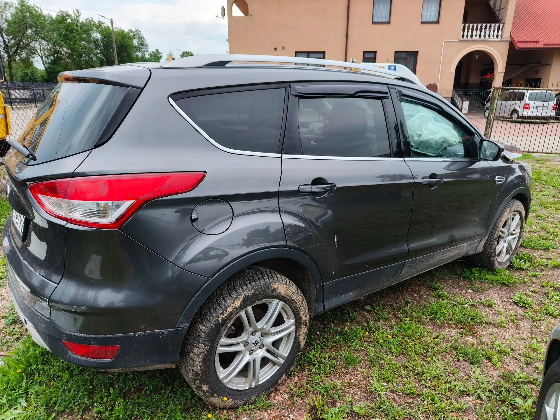 Szary samochód typu SUV zaparkowany na trawie. Pojazd jest widoczny z tyłu i z boku. Widoczne są uszkodzenia karoserii: wgniecenie na prawych tylnych drzwiach oraz rozbita przednia szyba pasażera, w której widoczna jest wypuszczona poduszka powietrzna. Samochód ma srebrne felgi aluminiowe i białe relingi dachowe. W tle znajduje się beżowy budynek z balkonem oraz inny, biały samochód.