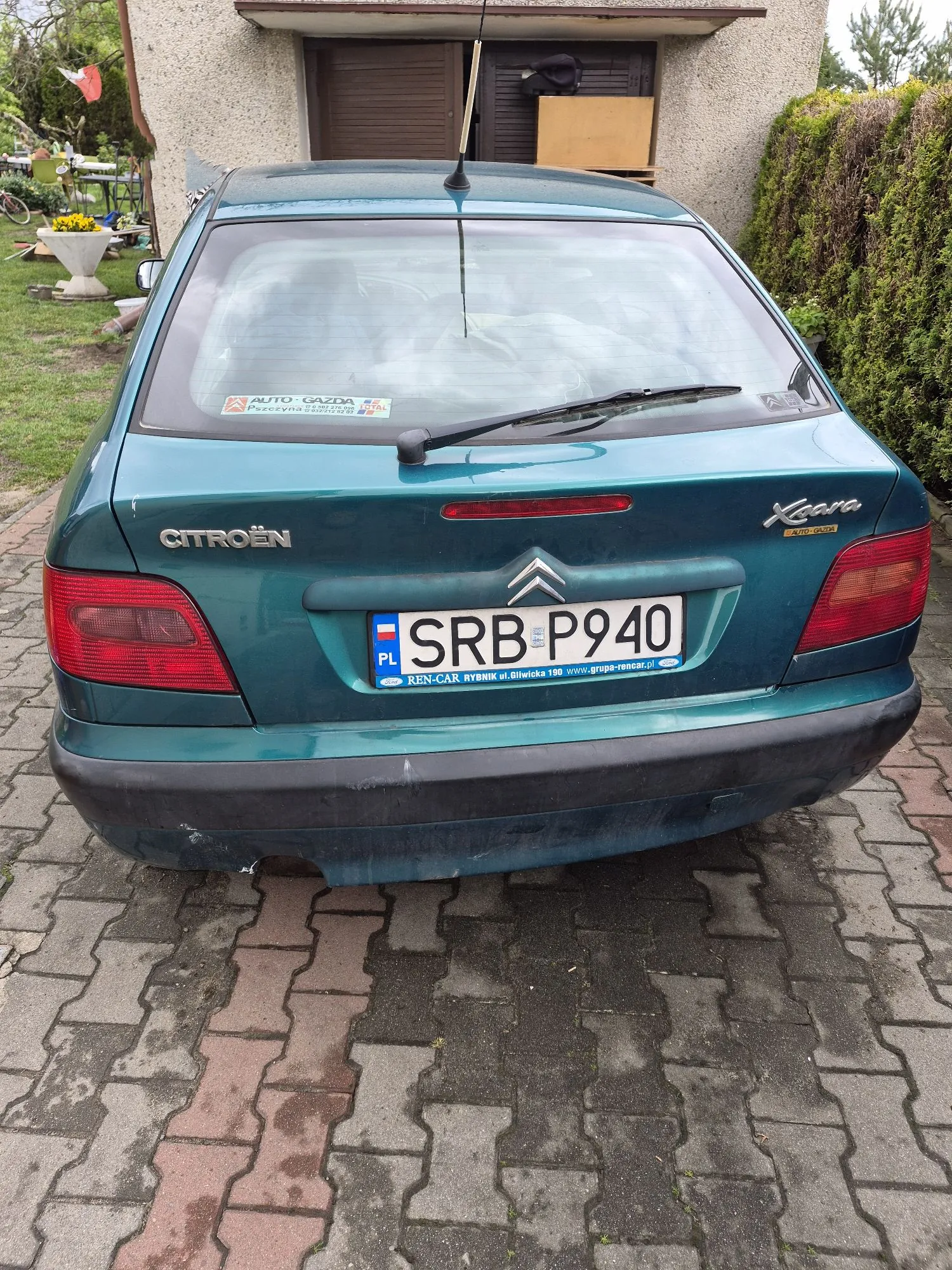 Tylny widok zielonego samochodu marki Citroën Xsara zaparkowanego na kostce brukowej przed domem. Na klapie bagażnika widnieje napis CITROËN, logo marki oraz oznaczenie modelu Xsara. Samochód posiada polską tablicę rejestracyjną o numerze SRB P940 z ramką dealera REN-CAR RYBNIK. Na tylnej szybie znajduje się naklejka AUTO-GAZDA.