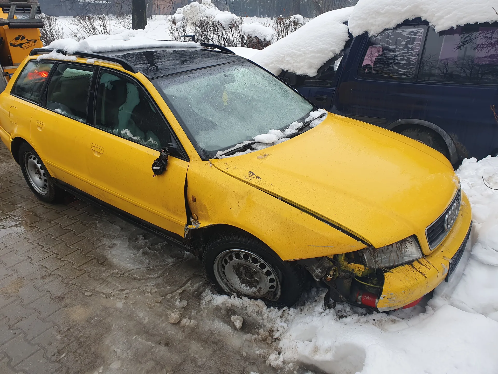 Zniszczony żółty samochód kombi z uszkodzonym przodem, pokryty śniegiem na dachu i przedniej szybie. Po lewej stronie widoczny niebieski samochód również pokryty śniegiem. Tło z zasnieżonymi krzewami i drzewami. Podłoga z kostki brukowej częściowo pokryta śniegiem i lodem. Na dachu żółtego samochodu znajduje się bagażnik dachowy.