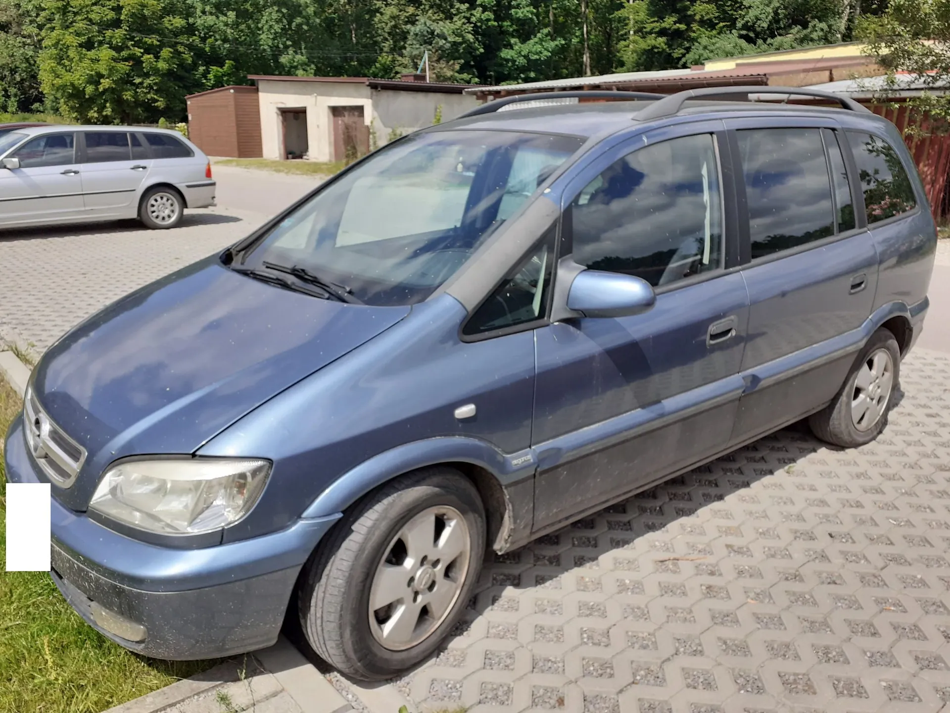 Niebieski samochód typu minivan zaparkowany na kostce brukowej. Pojazd jest widoczny z przodu i z boku. W tle znajduje się srebrny samochód osobowy, niskie budynki gospodarcze oraz gęste, zielone drzewa.
