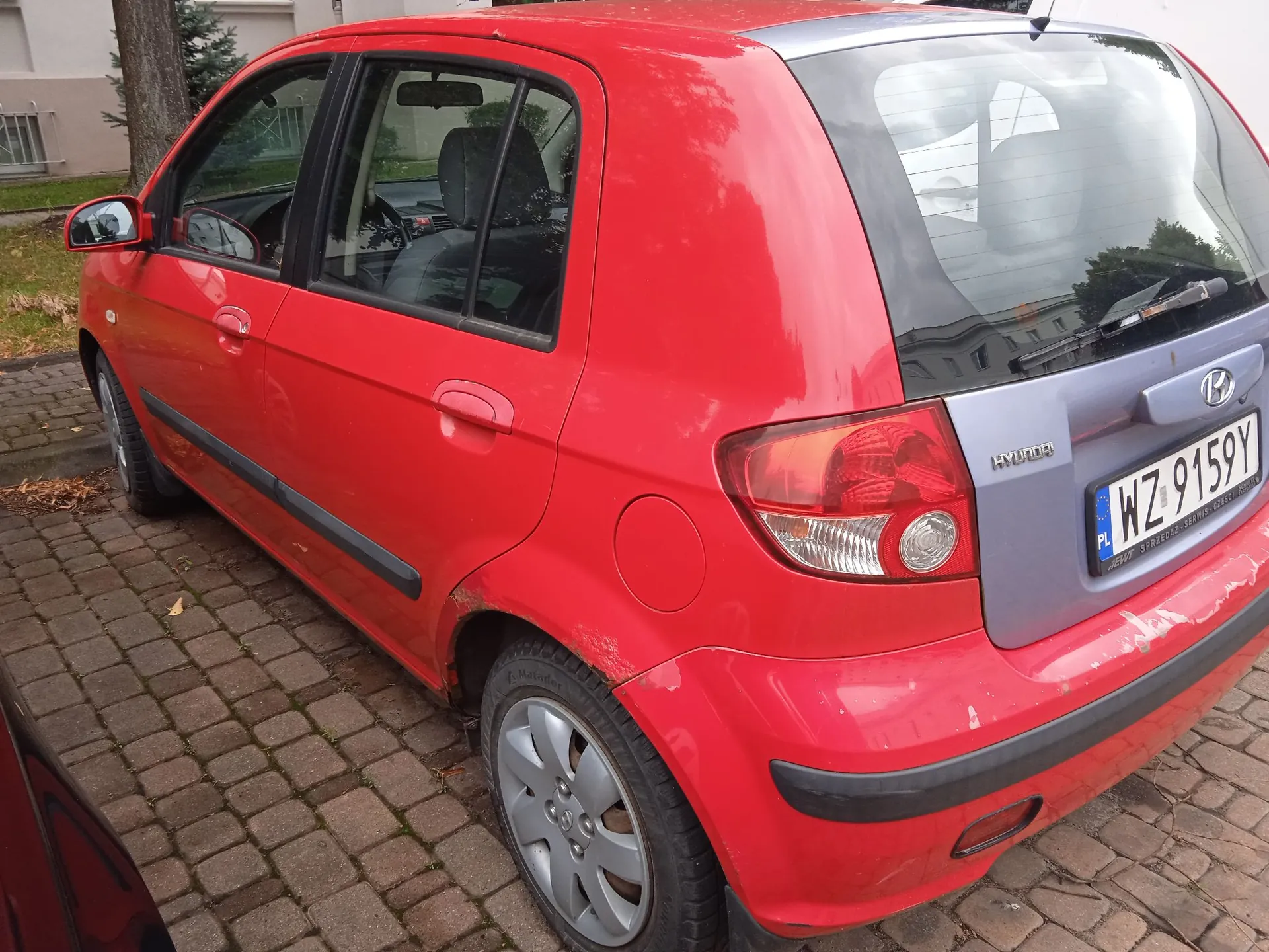 Czerwony samochód marki Hyundai z tyłu i z boku, zaparkowany na kostce brukowej. Pojazd posiada polską tablicę rejestracyjną o numerze WZ 9159Y. Na tylnym zderzaku i nad nadkolem tylnym lewym widoczne są uszkodzenia lakieru i rdza.