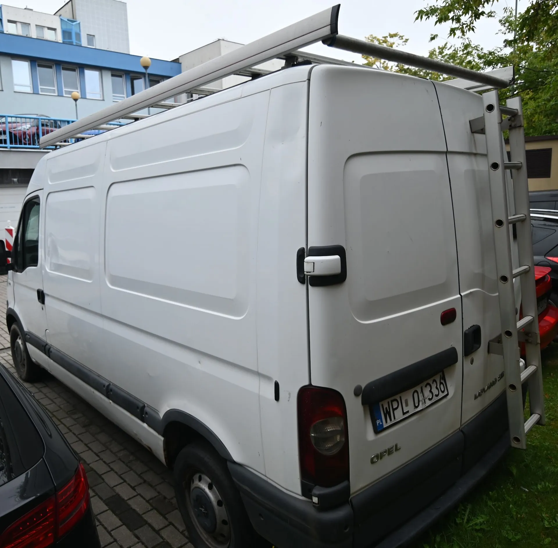 Biały furgon marki Opel zaparkowany na kostce brukowej. Pojazd posiada zamontowany na dachu srebrny bagażnik dachowy oraz drabinę z tyłu. Na tylnych drzwiach widoczna jest tablica rejestracyjna o numerze WPL 01336 oraz napis OPEL. W tle widoczny jest niebieski budynek biurowy oraz fragmenty innych samochodów.
