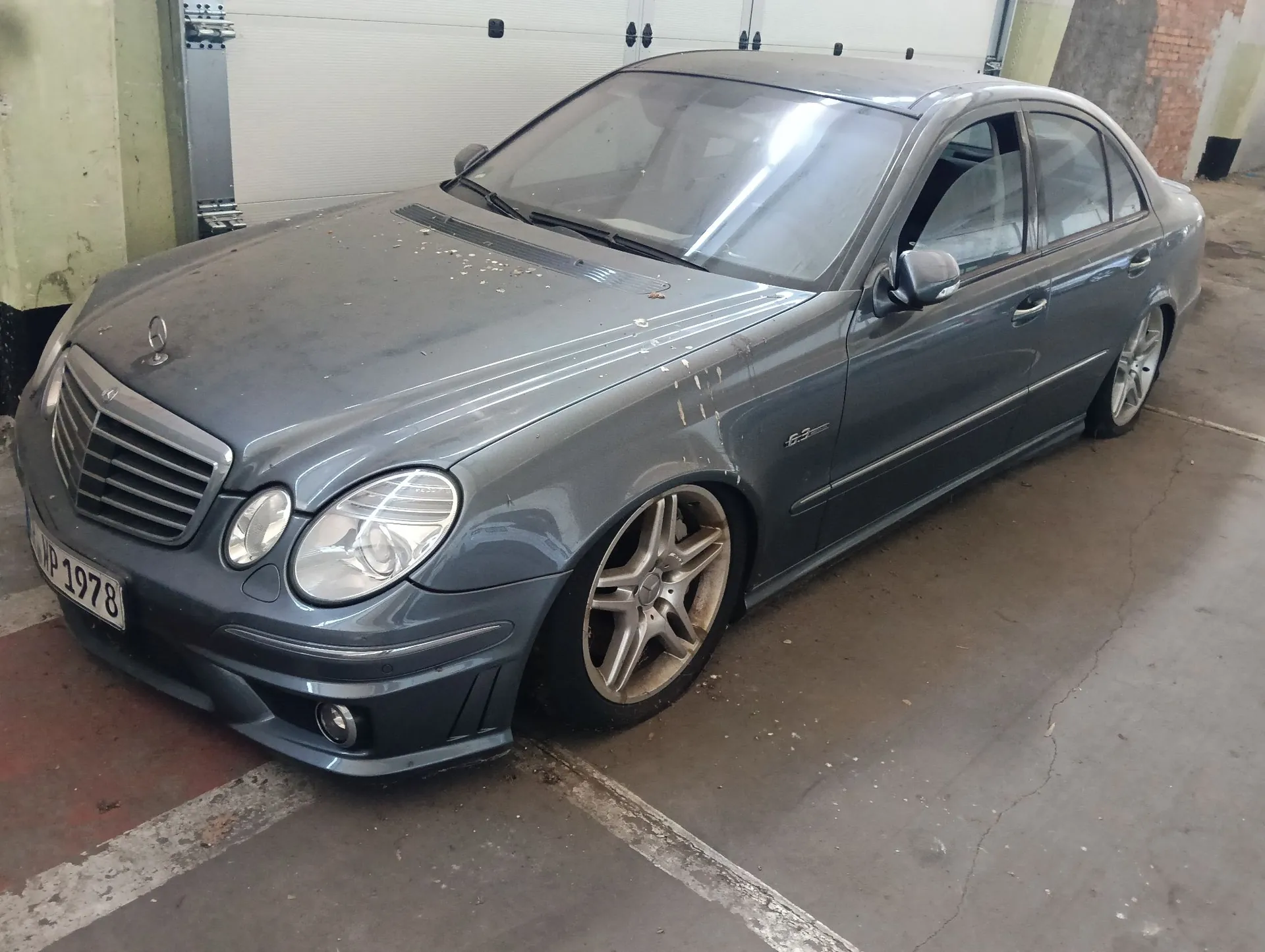 Srebrno-szary samochód osobowy marki Mercedes-Benz zaparkowany w garażu. Pojazd jest zakurzony, posiada felgi aluminiowe o pięciu ramionach oraz tablicę rejestracyjną o treści WP 1978. Na przednim błotniku widoczny jest napis E 63.