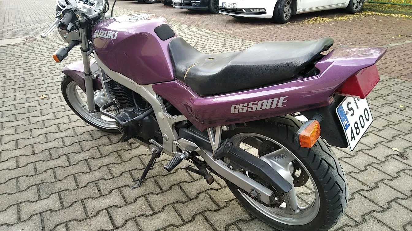 Fioletowy motocykl marki Suzuki, model GS500E, zaparkowany na kostce brukowej. Motocykl jest ustawiony bokiem, widoczna jest jego prawa strona, czarne siedzenie oraz tablica rejestracyjna o numerze SI 4800. W tle widoczne są zaparkowane samochody.