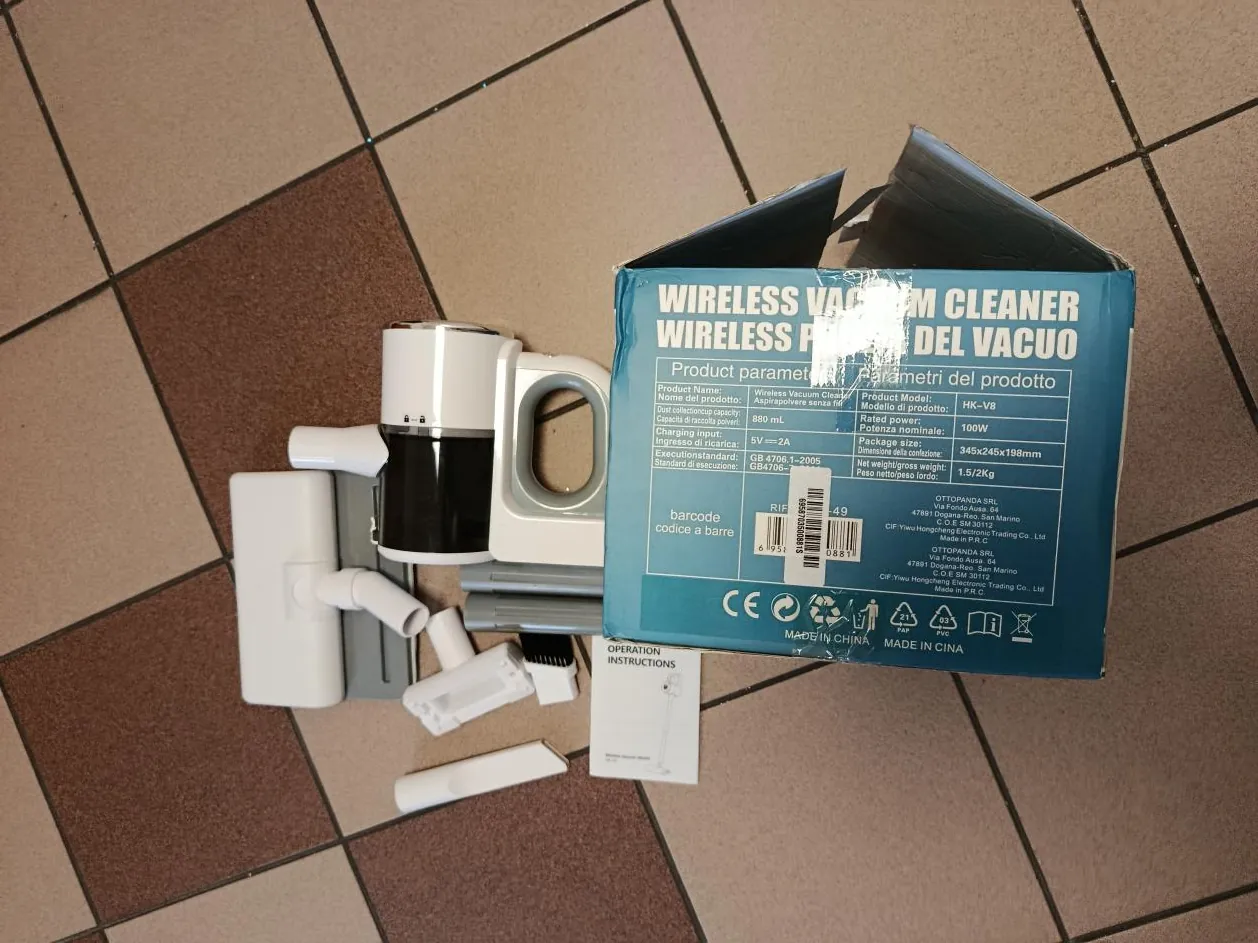 Zdjęcie przedstawia niebieskie pudełko z napisem WIRELESS VACUUM CLEANER oraz parametrami produktu w języku włoskim, w tym frazą DEL VACUO. Na pudełku widoczne są symbole CE, kod kreskowy, informacja MADE IN CHINA oraz dane techniczne. Obok pudełka leżą części odkurzacza: biało-czarna jednostka główna, różne nasadki, szczotka i instrukcja. Tło stanowi podłoga z brązowo-beżowymi płytkami. Tekst na pudełku jest odwrócony do góry nogami.