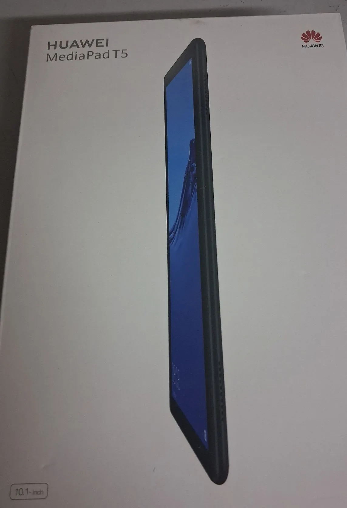 Białe opakowanie tabletu Huawei MediaPad T5. Na pudełku widnieje napis 'HUAWEI MediaPad T5' w lewym górnym rogu oraz logo Huawei w prawym górnym rogu. W centrum znajduje się zdjęcie tabletu pokazanego z profilu, z włączonym niebieskim ekranem. W lewym dolnym rogu widnieje napis '10.1-inch'.