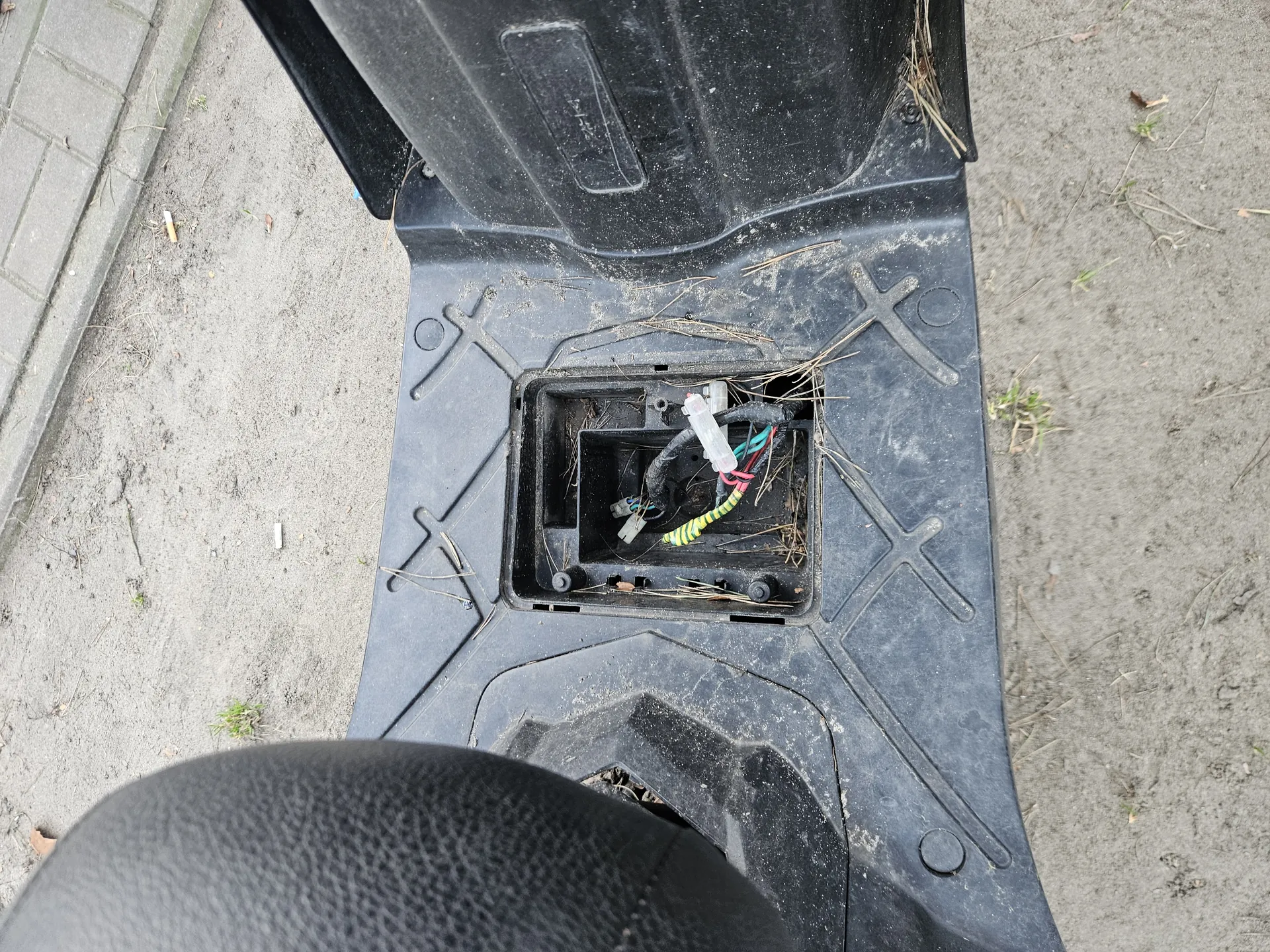 Czarna plastikowa konstrukcja z otwartym przedziałem zawierającym przewody elektryczne w kolorach zielonym, żółtym, czerwonym i czarnym oraz biały złącznik. Na powierzchni widoczny brud, sucha trawa i drobne śmieci. W tle betonowa powierzchnia z fragmentami trawy i chodnikiem z płytek. W dolnej części obrazu fragment czarnej, teksturowanej powierzchni.