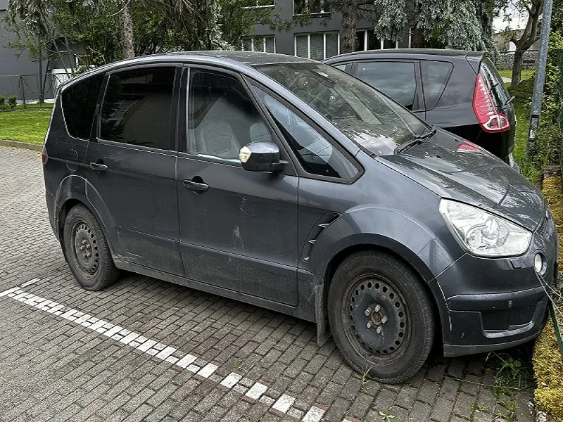 Ciemnoszary samochód osobowy typu minivan zaparkowany na brukowanym placu parkingowym. Pojazd ma zabrudzone boki i brakującą nakładkę na przednim prawym kole. W tle widoczny czarny samochód, zielona trawa, drzewa oraz budynek z oknami. Na nawierzchni białe linie parkingu.