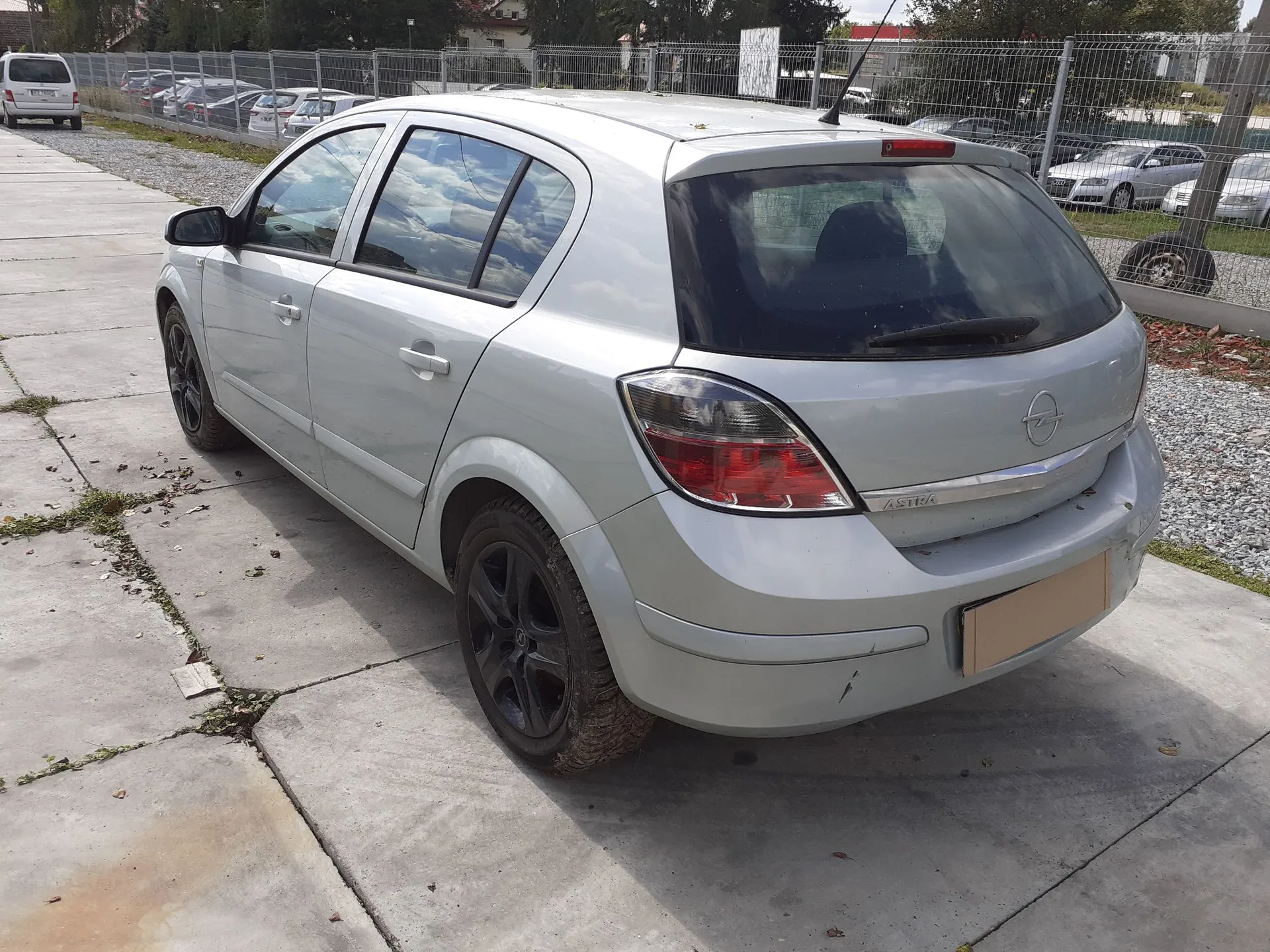 Srebrny samochód marki Opel Astra w nadwoziu hatchback, zaparkowany na betonowym podjeździe. Auto jest widoczne z tyłu i z boku, posiada czarne felgi aluminiowe. W tle znajduje się ogrodzenie z siatki, inne zaparkowane samochody oraz drzewa.