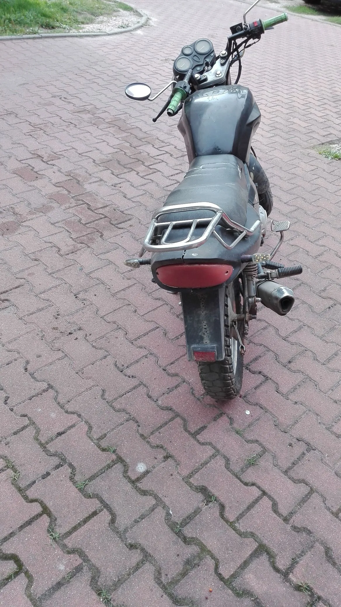 Czarny motocykl zaparkowany tyłem do widza na kostce brukowej. Pojazd posiada srebrny bagażnik z tyłu, czerwony odblask oraz widoczną rurę wydechową po prawej stronie. Kierownica ma zielone manetki.