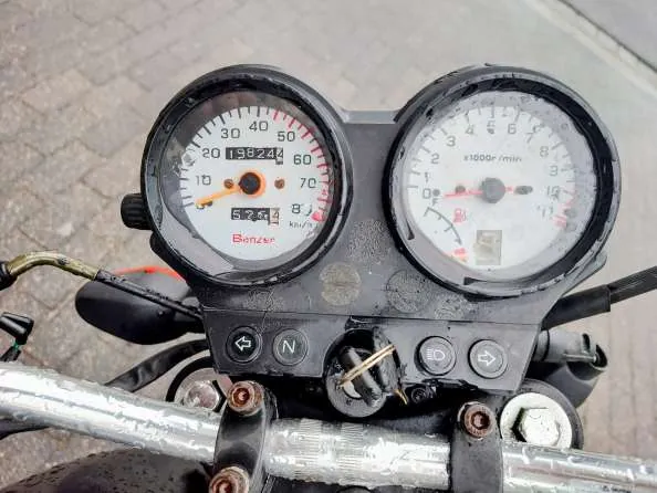 Zbliżenie na deskę rozdzielczą motocykla z dwoma głównymi wskaźnikami. Po lewej stronie prędkościomierz z zakresem 0-70 km/h, czerwoną strefą od 50 km/h, cyfrowym licznikiem przebiegu 19824 i dodatkowym wyświetlaczem 52.5. Poniżej widoczny napis 'Benzler' w czerwonej kolorystyce. Po prawej tachometr z zakresem 0-11 (x1000r/min). Poniżej wskaźników umieszczone są przyciski oraz wskaźnik biegu 'N'. Widoczne metalowe kierownice z zardzewiałymi śrubami. Tło stanowi brukowana powierzchnia.