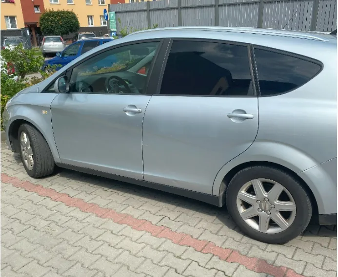 Srebrny samochód typu hatchback zaparkowany bokiem na kostce brukowej. W tle widoczny jest budynek mieszkalny, zielone krzewy oraz szary płot.