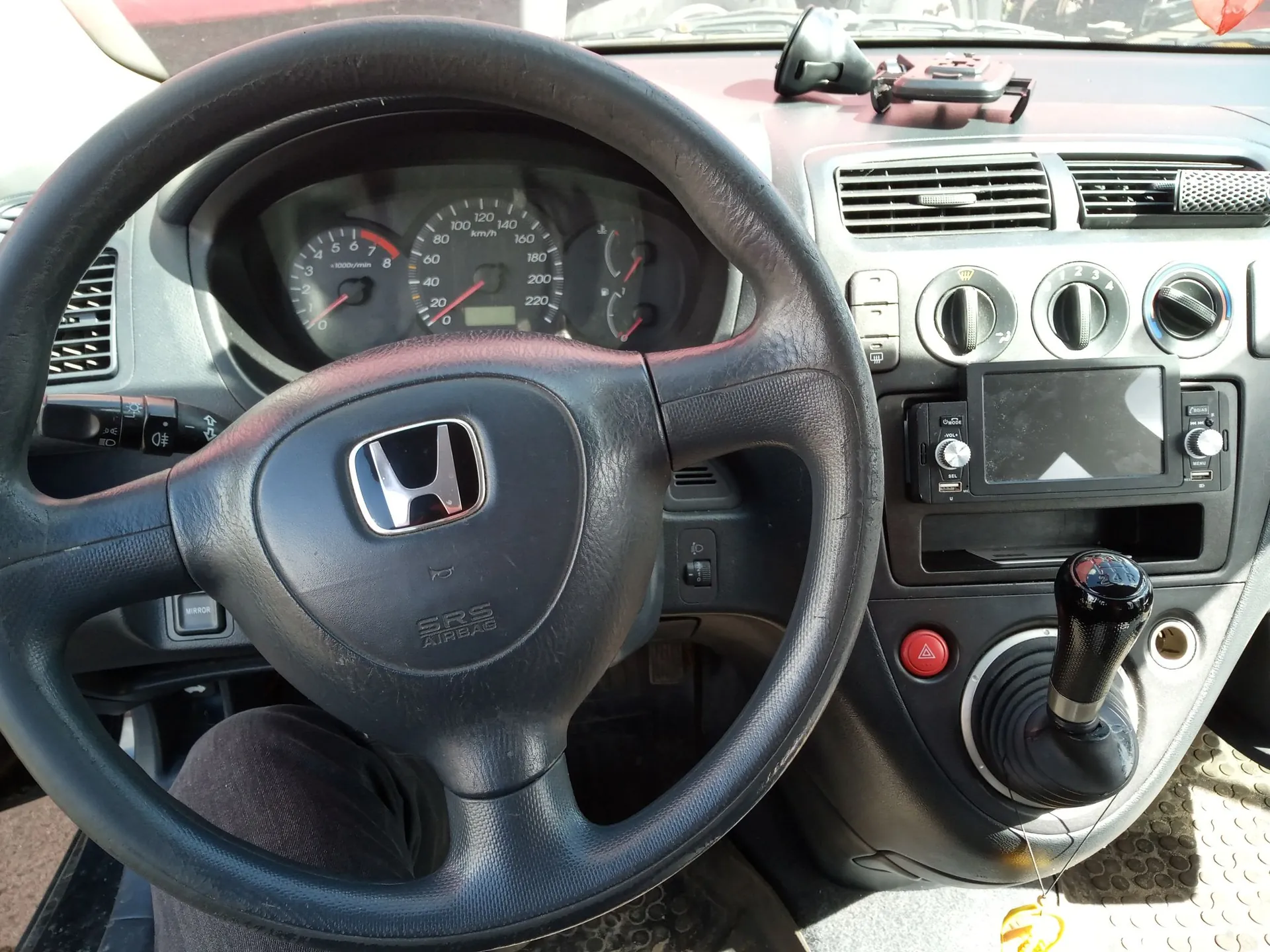 Wnętrze samochodu marki Honda, widok z perspektywy kierowcy. Na pierwszym planie znajduje się czarna, skórzana kierownica z logo Hondy w centrum i napisem SRS AIRBAG. W tle widoczna jest deska rozdzielcza z analogowymi wskaźnikami prędkości i obrotów, centralna konsola z pokrętłami klimatyzacji, ekranem radia oraz lewarkiem zmiany biegów z czarną gałką. Po prawej stronie znajduje się czerwony przycisk.