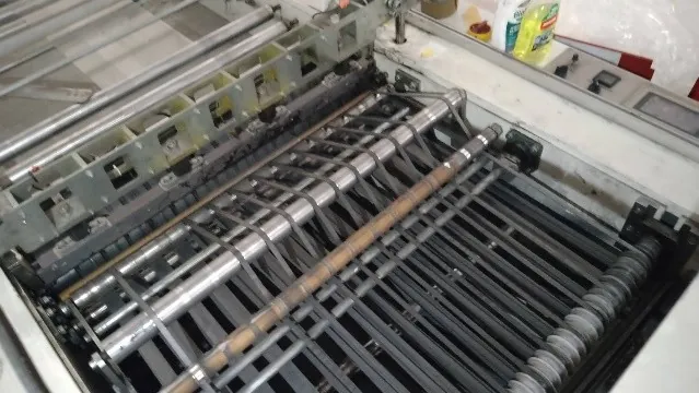 Zbliżenie na wnętrze maszyny przemysłowej z systemem metalowych wałków i prowadnic. Widoczne są liczne srebrne rurki ułożone równolegle do siebie oraz metalowa konstrukcja z otworami. W prawym górnym rogu znajduje się biała powierzchnia z butelką żółtego płynu czyszczącego i panelem sterowania.