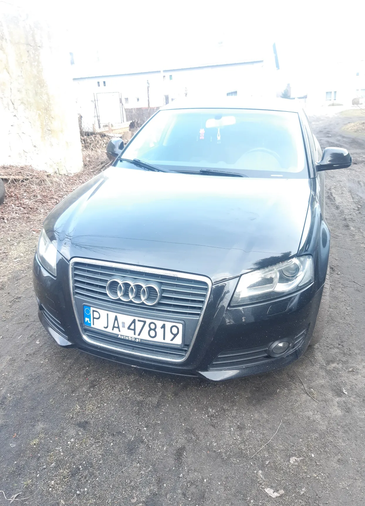 Czarny samochód marki Audi z tablicą rejestracyjną PJA 47819. Widoczne przednie światła, logo Audi na masce oraz podłoże z żwiru. W tle fragment budynku i sucha trawa.