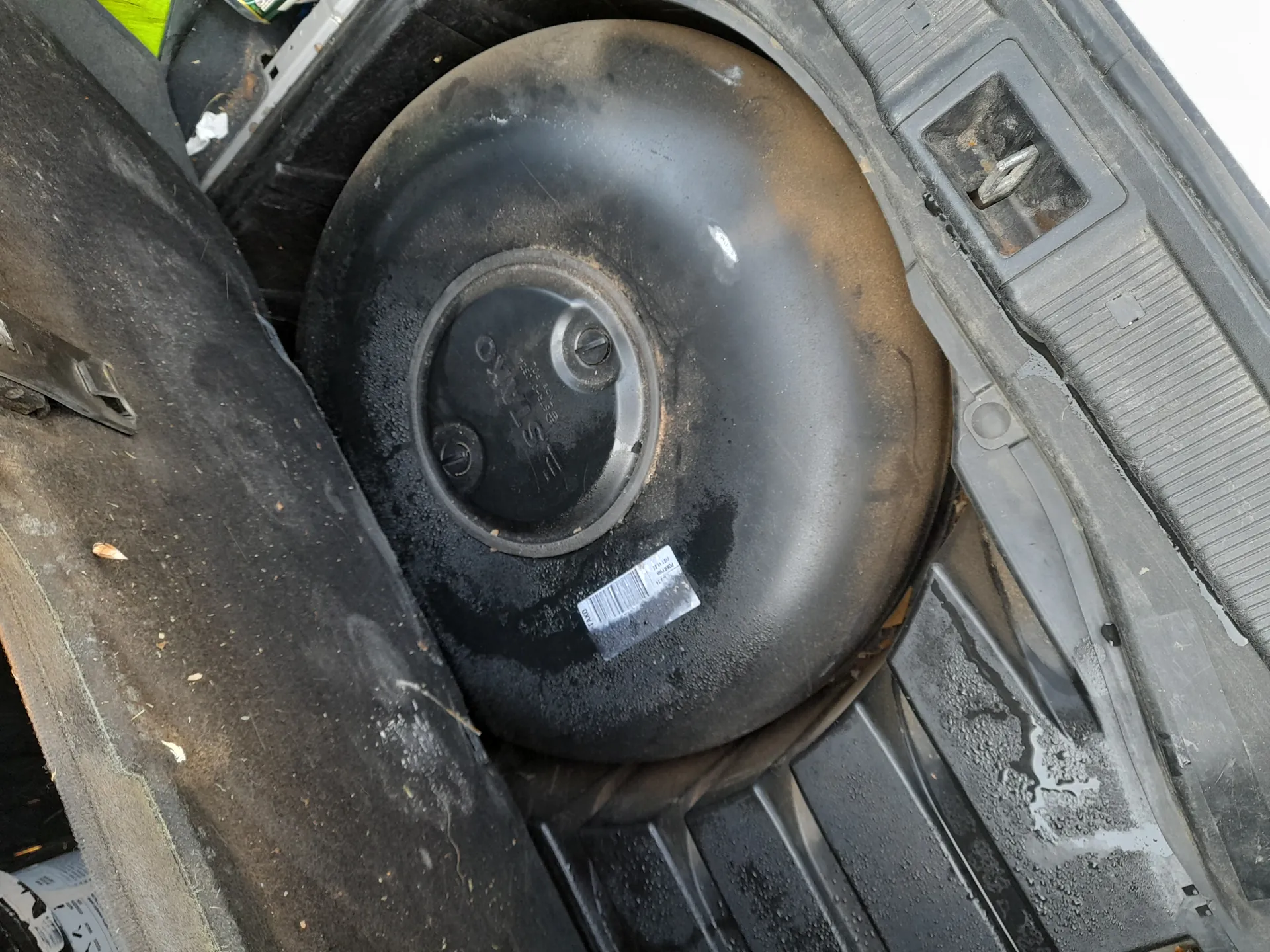 Zbliżenie na zapasowe koło umieszczone w bagażniku samochodu. Czarne koło z widocznymi śladami brudu i wilgoci, z naklejką zawierającą kod kreskowy na powierzchni. W centrum koła znajduje się okrągła pokrywa z napisem 'Spare' oraz dodatkowymi oznaczeniami. Wokół koła widoczne są elementy wnętrza bagażnika, w tym czarne plastikowe panele z zabrudzeniami i resztkami materiału.