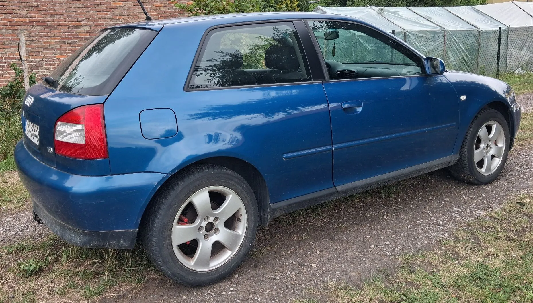 Niebieski samochód osobowy marki Audi w nadwoziu hatchback, zaparkowany bokiem na szutrowej drodze. W tle widoczny jest ceglany mur oraz biała konstrukcja szklarni. Samochód ma srebrne felgi aluminiowe z pięcioma ramionami.