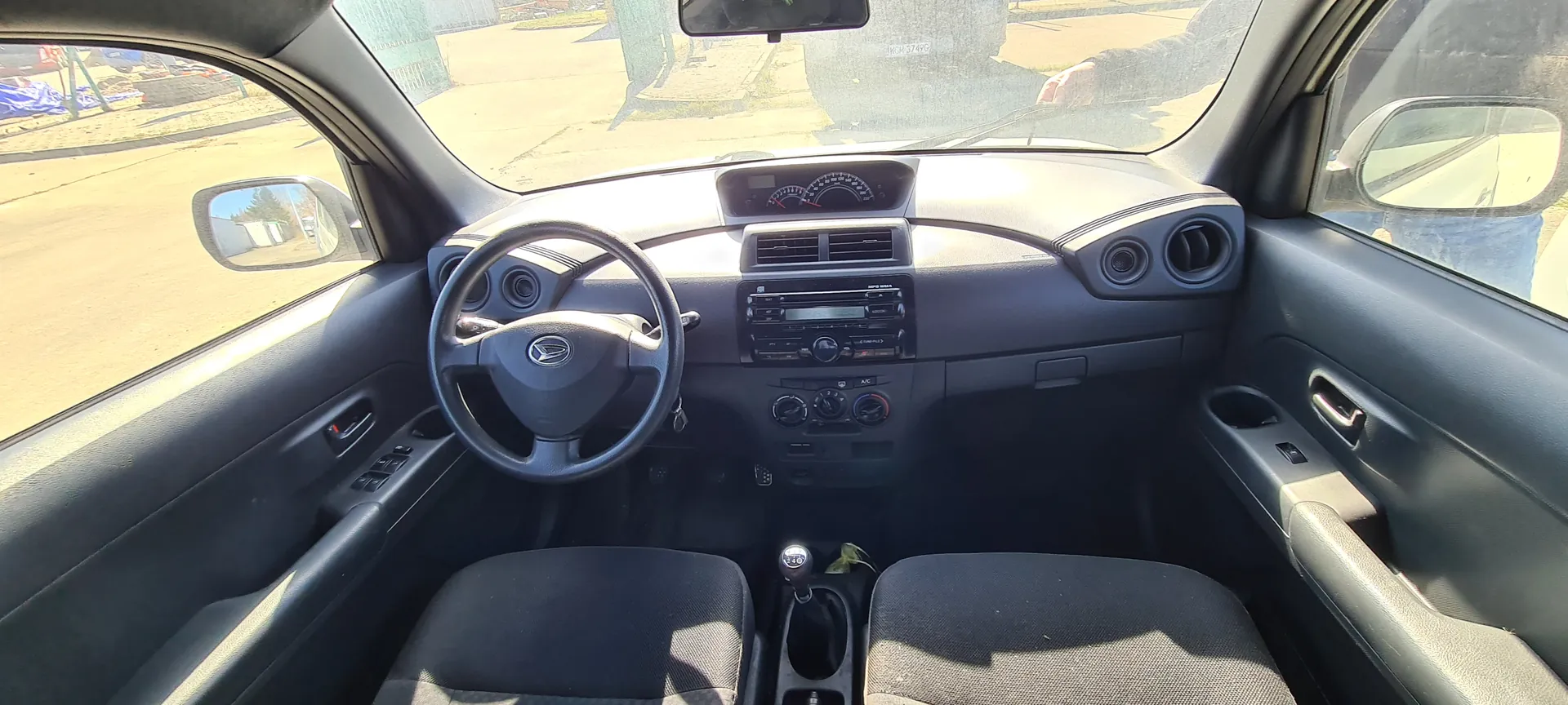 Wnętrze samochodu widziane z tyłu na przód. Kierownica z logo Daihatsu po lewej stronie, czarna deska rozdzielcza z licznikami, systemem audio i wentylacją. Czarne siedzenia, dźwignia biegów między nimi. Widoczne lusterka boczne i przednie szyby z widokiem na ulicę i budynki na zewnątrz. Wnętrze w odcieniach czerni i szarości, oświetlenie dziennego światła.