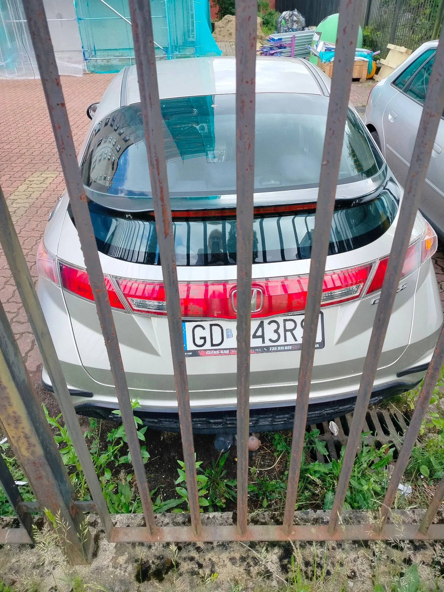 Zdjęcie tyłu srebrnego samochodu marki Honda, wykonane przez metalowy płot z pionowych prętów. Widoczna jest tablica rejestracyjna o numerze GD 43RS. Samochód zaparkowany jest na kostce brukowej, a pod nim widoczna jest trawa i roślinność.