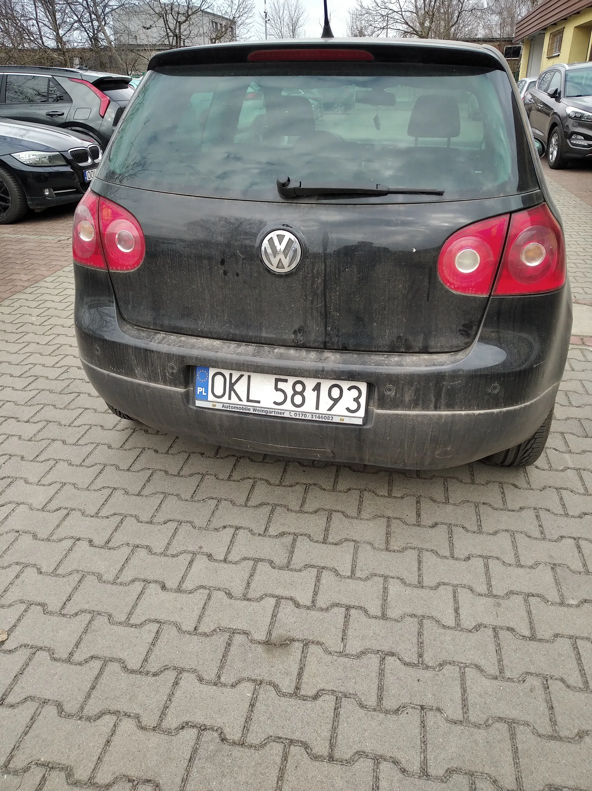 Tył czarnego samochodu marki Volkswagen z brudną karoserią, z widocznym logo VW na bagażniku. Tablica rejestracyjna z białym tłem i czarnym napisem OKL 58193, z niebieskim pasem po lewej stronie z literą PL oraz napisem Automobile Weingartner i numerem telefonu 0170/3146082. Czerwone światła tylne, czyste szyby i wycieraczka na tylnej szybie. Samochód zaparkowany na brukowanej powierzchni, w tle inne pojazdy (m.in. czarny BMW) oraz budynki i drzewa bez liści.
