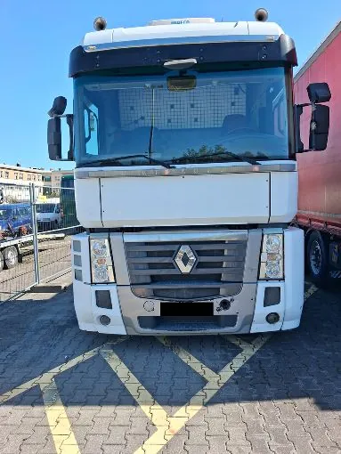 Widok z przodu białej ciężarówki marki Renault zaparkowanej na kostce brukowej z żółtymi liniami wyznaczającymi strefę. Pojazd posiada duże przednie szyby, lusterka boczne oraz charakterystyczny grill z logo Renault na środku. Po prawej stronie widoczny jest fragment czerwonej naczepy.
