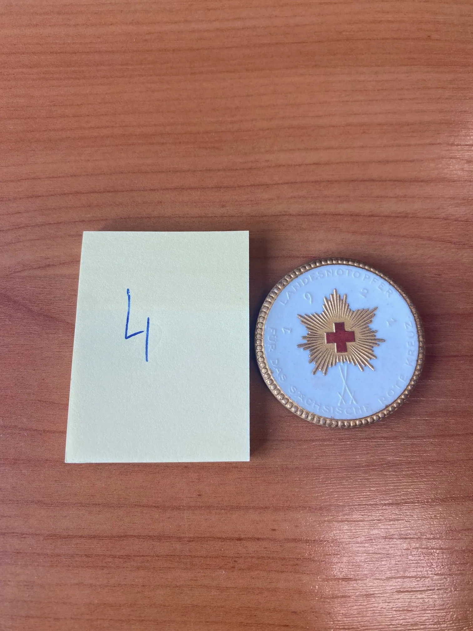 Na drewnianym stole leży prostokątny, jasnozielony kawałek papieru z niebieską cyfrą 4 oraz okrągły medal. Medal ma jasnoniebieskie tło, czerwony krzyż w centrum, otoczony złotym promienistym wzorem. Wokół medalu widoczny jest napis: LANDESNOTOPFER 1921 FÜR DAS SÄCHSISCHE ROTE KREUZ. Brzeg medalu zdobiony jest złotą, kropkowaną obramówką.