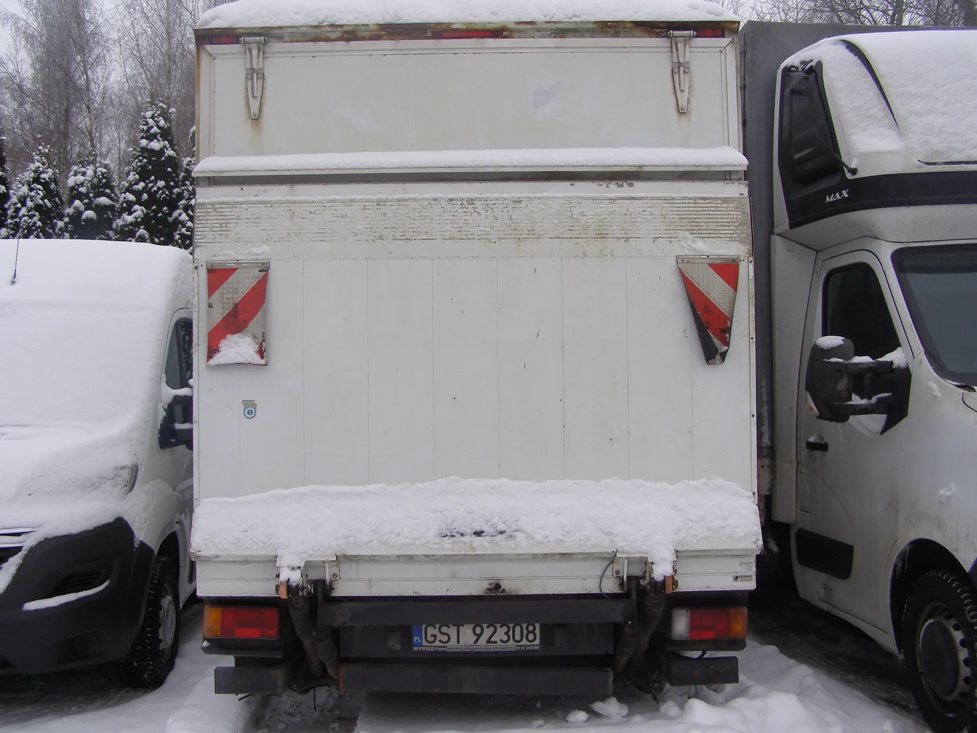 Tył białej ciężarówki z tablicą rejestracyjną GST 92308, pokryty śniegiem. Czerwono-białe paski ostrzegawcze na drzwiach. Po lewej furgonetka, po prawej ciężarówka z napisem MAX. Śnieg na dachach, ziemi i drzewach w tle. Widoczne ślady zużycia na nadwoziu.