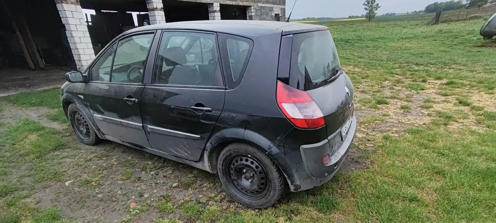 Czarny samochód osobowy typu hatchback, zabrudzony błotem, zaparkowany na trawiastym i błotnistym terenie. W tle widoczny niedokończony budynek z białymi blokami i otwartymi przestrzeniami, a dalej rozległe pole z zieloną trawą i pojedynczym drzewem. Niebo jest pochmurne. Samochód ma czarne felgi i czerwone światła tylnego. Na tablicy rejestracyjnej widoczne są nieczytelne znaki.