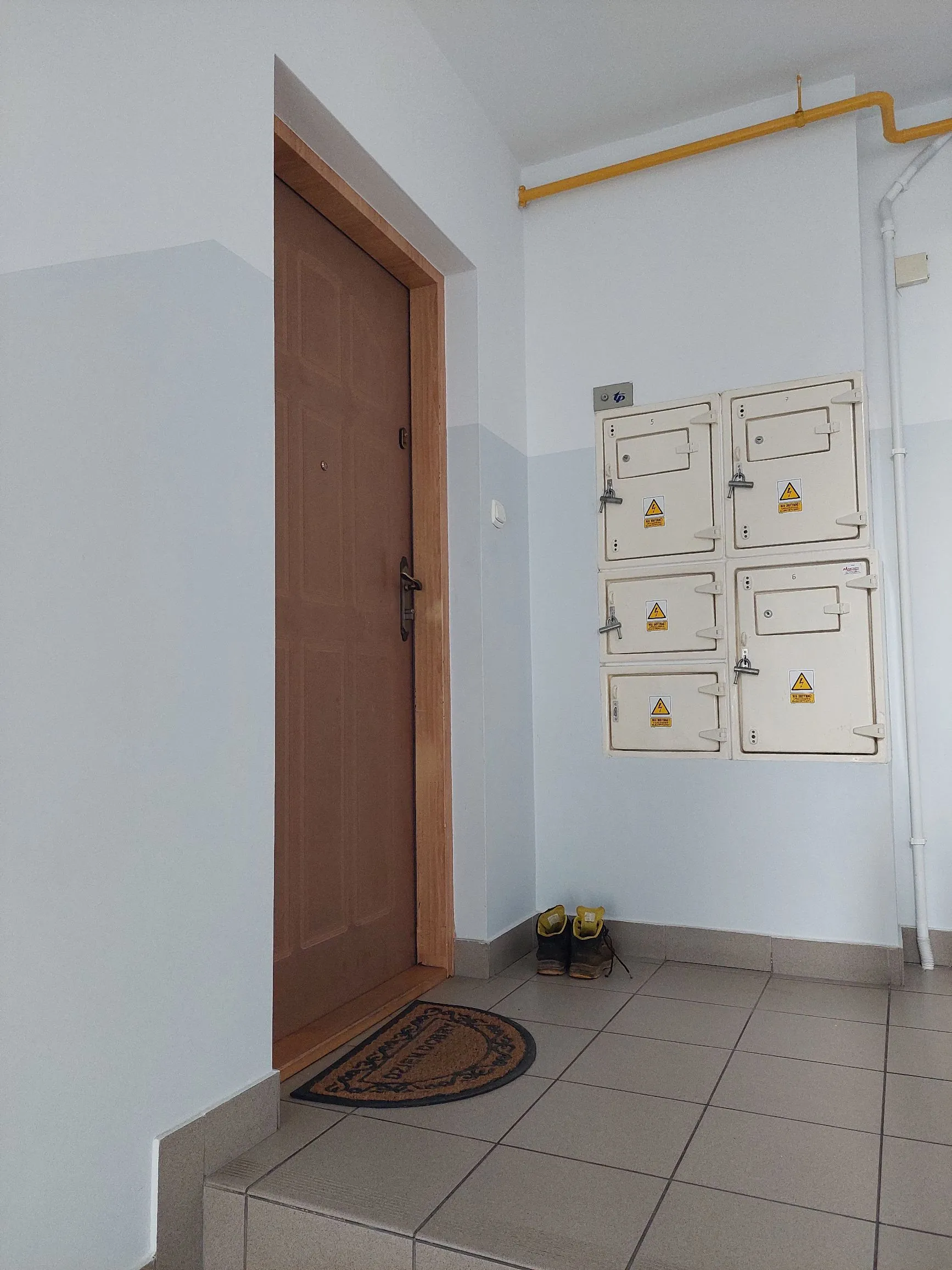Wnętrze korytarza z jasnymi ścianami, których dolna część jest pomalowana na jasnoniebieski kolor. Po lewej stronie znajduje się brązowe, drewniane drzwi w drewnianej ościeżnicy. Przed drzwiami leży ciemna wycieraczka w kształcie owalu. Na podłodze z jasnych płytek ceramicznych stoją żółto-czarne buty robocze. Na prawej ścianie zamontowane są cztery białe skrzynki elektryczne z żółtymi ostrzeżeniami przed wysokim napięciem. Nad skrzynkami biegnie żółta rura gazowa.