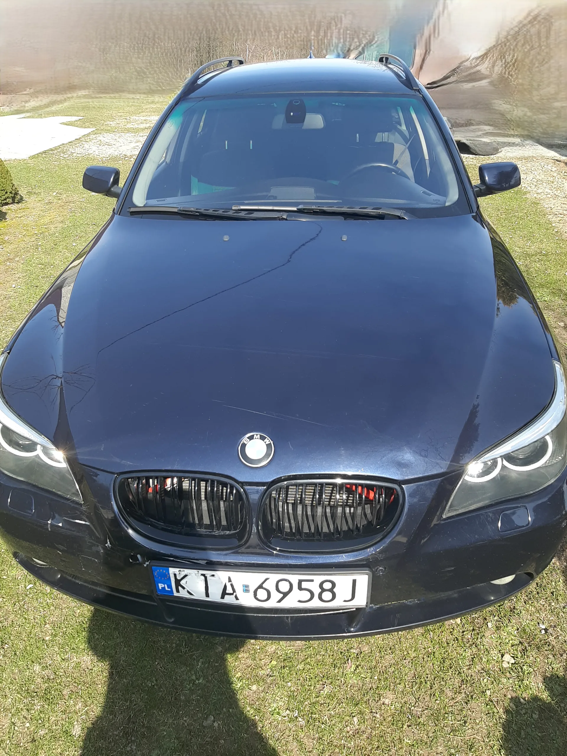 Ciemnoniebieski samochód marki BMW z przodu, z widocznym logo na masce i charakterystyczną podwójną atrapą chłodnicy. Tablica rejestracyjna z białym tłem i czarnym napisem KTA 6958 J, z niebieskim pasem po lewej stronie z napisem PL. Samochód stoi na trawie z widocznymi plamami żwiru, na tle rozmytego budynku i suchych krzewów. Światła przednie są włączone, a na masce widoczne są drobne zadrapania. W tle widoczne są cienie i fragmenty otoczenia.