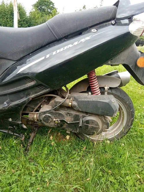 Zbliżenie na tylną część czarnego skutera zaparkowanego na zielonej trawie. Na boku obudowy widnieje biały napis 'FORMAX'. Widoczny jest czerwony amortyzator, tylne koło, silnik oraz czarna kanapa.