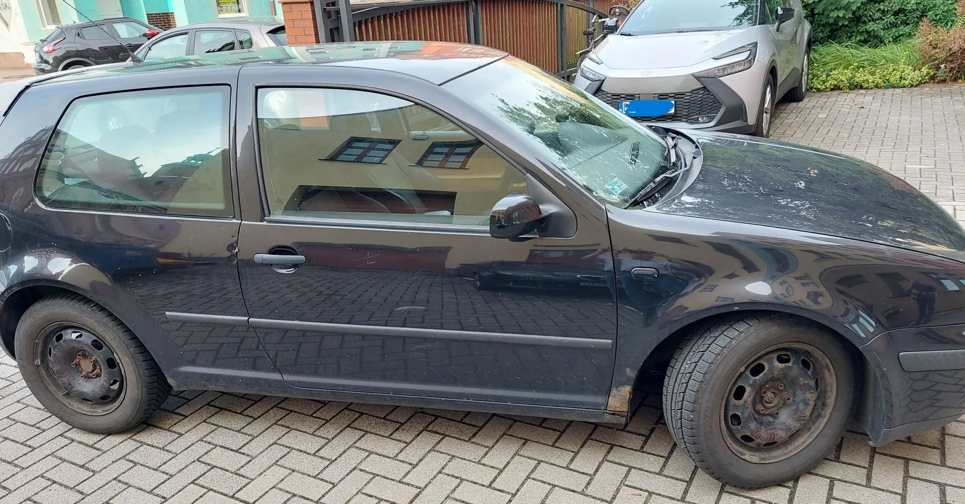 Czarny, trzydrzwiowy samochód osobowy zaparkowany bokiem na kostce brukowej. W tle widoczne są inne zaparkowane samochody, w tym srebrny SUV, oraz fragmenty budynków i zieleni.