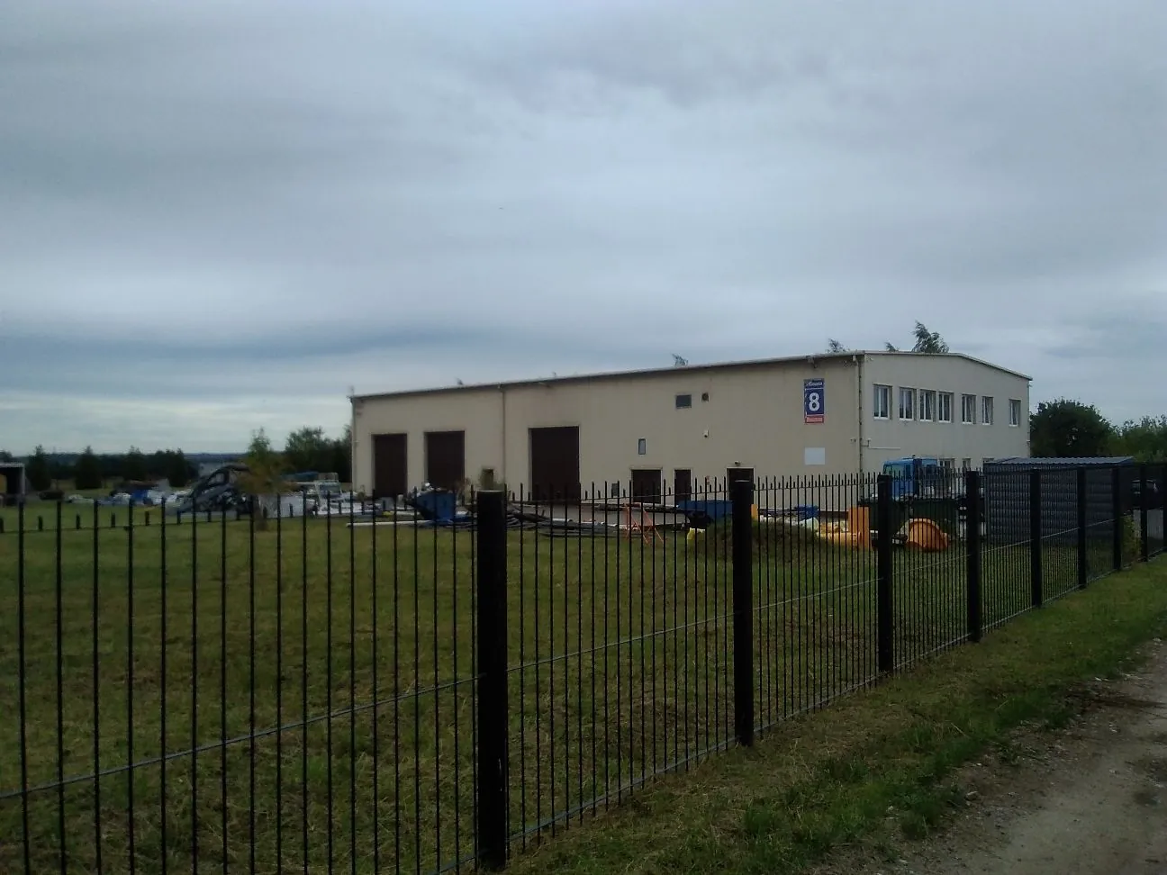 Zdjęcie przedstawia jednokondygnacyjny, jasny budynek przemysłowy lub magazynowy z kilkoma bramami wjazdowymi i oknami po prawej stronie. Na ścianie budynku widnieje niebieska tabliczka z białą cyfrą 8. Przed budynkiem znajduje się teren z trawą, ogrodzony czarnym, metalowym płotem z prętów. W tle widać pochmurne, szare niebo.