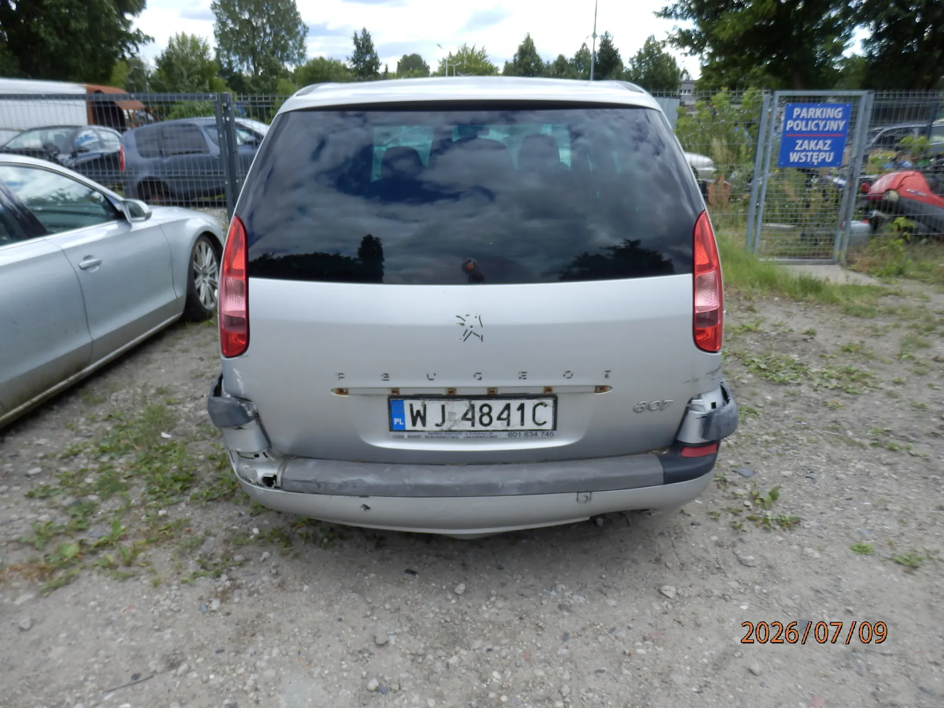 Srebrny samochód marki Peugeot 607 widziany od tyłu, zaparkowany na piaszczystym placu. Pojazd ma uszkodzony tylny zderzak po lewej stronie. Tablica rejestracyjna ma numer WJ 4841C. W tle widoczne są inne samochody, ogrodzenie z siatki oraz niebieska tabliczka z napisem: 'PARKING POLICYJNY ZAKAZ WSTĘPU'. W prawym dolnym rogu zdjęcia znajduje się data 2026/07/09.