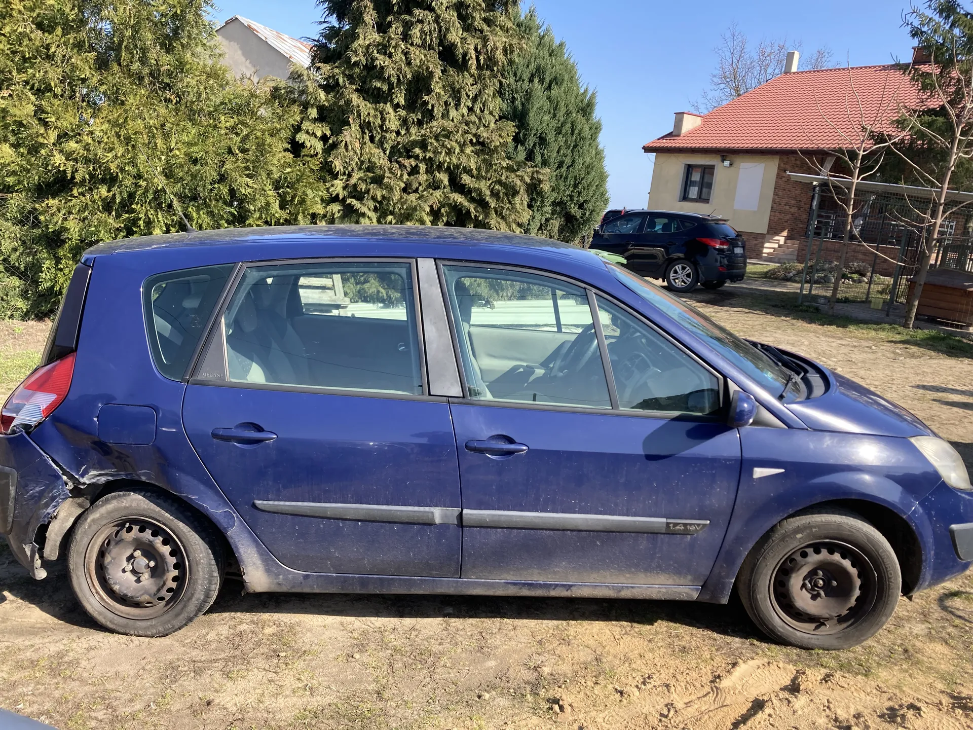 Niebieski samochód osobowy typu hatchback z widocznym uszkodzeniem na lewym tylnym zderzaku i brudną karoserią. Na boku pojazdu widoczny napis '1.4 16V'. Samochód stoi na suchym, ziemistym terenie z rzadką trawą. W tle dom z czerwoną dachówką, czarny samochód osobowy, zielone krzewy i drzewa oraz jasne niebo. Koła pojazdu mają rdzewne felgi.