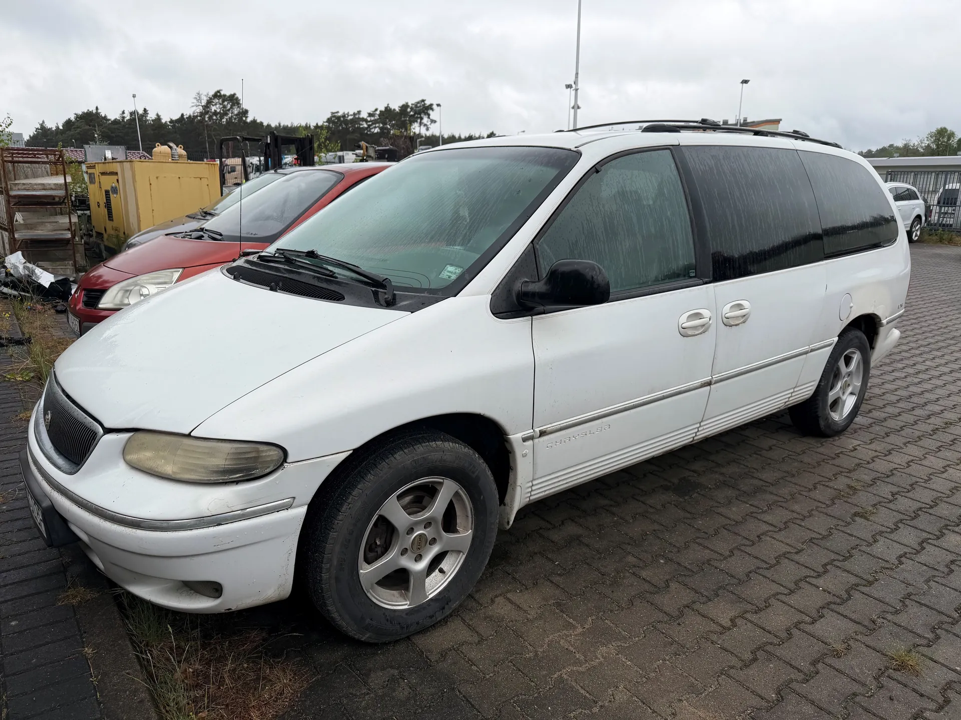 Biały minivan marki Chrysler Voyager zaparkowany na brukowanej powierzchni. Pojazd posiada czarne przyciemniane szyby, dachowy bagażnik i srebrne felgi. W tle widoczne są czerwone i szare samochody, żółty kontener, drzewa oraz przemysłowe konstrukcje. Niebo jest zachmurzone. Na boku minivana widoczny napis "CHRYSLER".