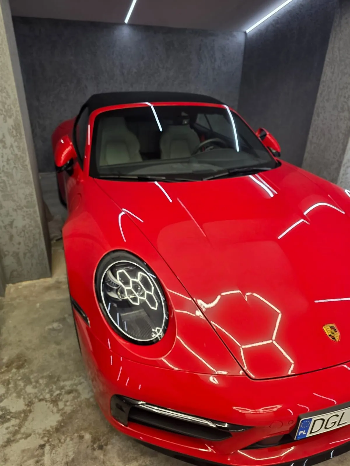 Czerwony samochód sportowy Porsche typu kabriolet zaparkowany w garażu. Pojazd ma czarny dach, jasne siedzenia, reflektory z geometrycznym wzorem i tablicę rejestracyjną z napisem PL DGL. W tle betonowe ściany i sufit z oświetleniem LED. Na masce widoczny emblemat marki.