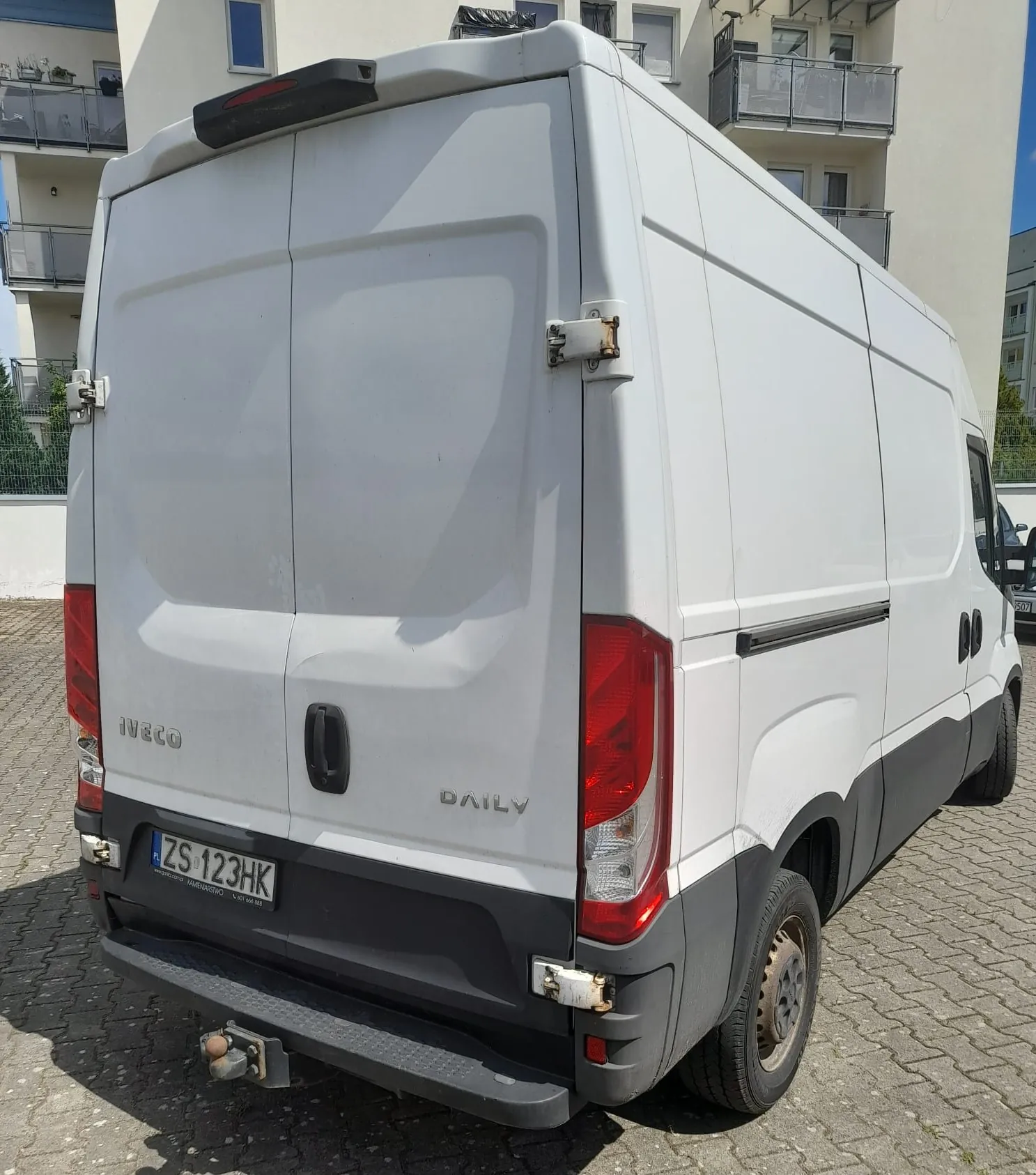Biały furgon marki Iveco Daily zaparkowany na kostce brukowej przed białym budynkiem mieszkalnym. Widoczny jest tył i prawy bok pojazdu. Na tylnych drzwiach znajduje się napis 'IVECO' oraz 'DAILY'. Tablica rejestracyjna ma numer 'ZS 123HK'. Pojazd posiada hak holowniczy.